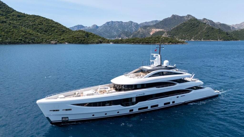 A luxurious white superyacht glides through the calm, blue waters of a scenic bay, with lush green hills in the background.