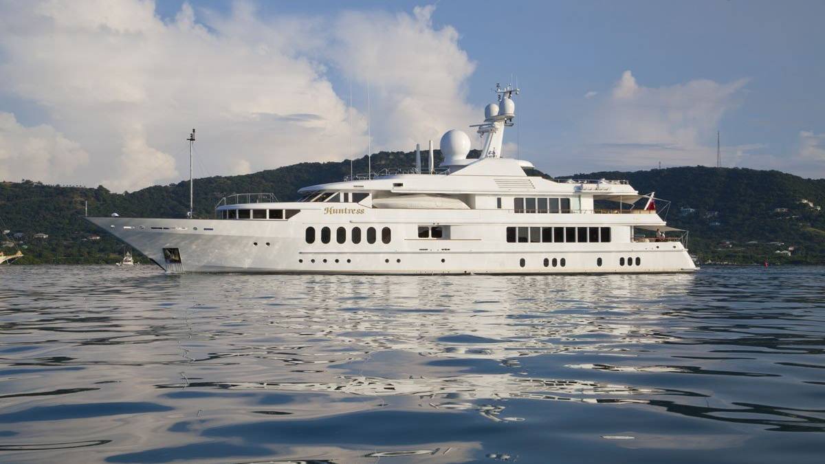 The luxurious yacht Huntress glides serenely across the calm, reflective waters under a bright blue sky.