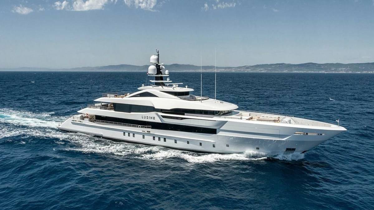 A large, luxurious white yacht sailing across the deep blue ocean, with distant hills or mountains visible in the background under a partly cloudy sky.
