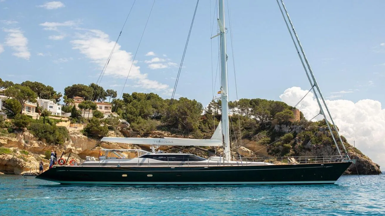 A sleek, dark green yacht floats on turquoise water in the foreground, with a lush, tree-covered hillside dotted with white buildings in the background.