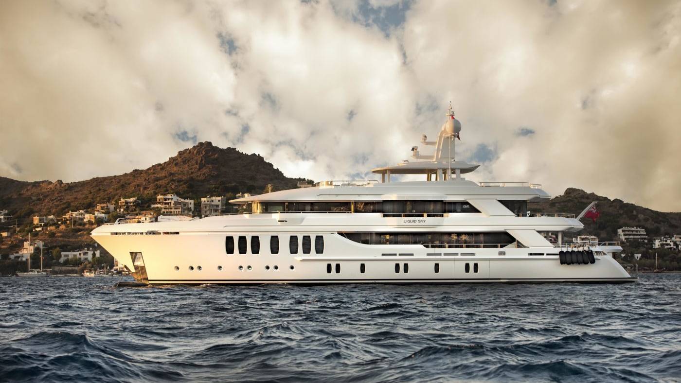 A luxurious white yacht named "Liquid Sky" cruises on choppy blue waters with a hillside village in the background under a dramatic sky.