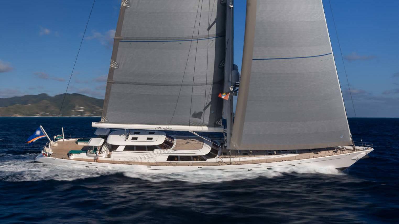 A large white sailboat with full sails glides through dark blue water, with green, hilly islands visible in the hazy background under a bright blue sky.