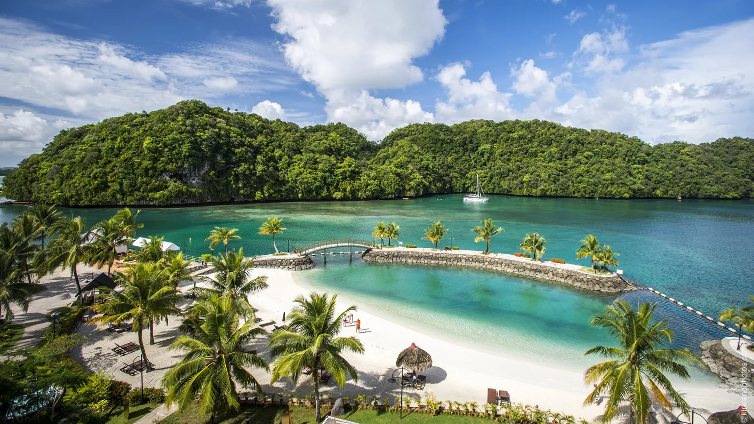Palm trees line a white sand beach and a calm turquoise lagoon in the foreground, while lush green islands and a sailboat dot the background under a cloudy blue sky.