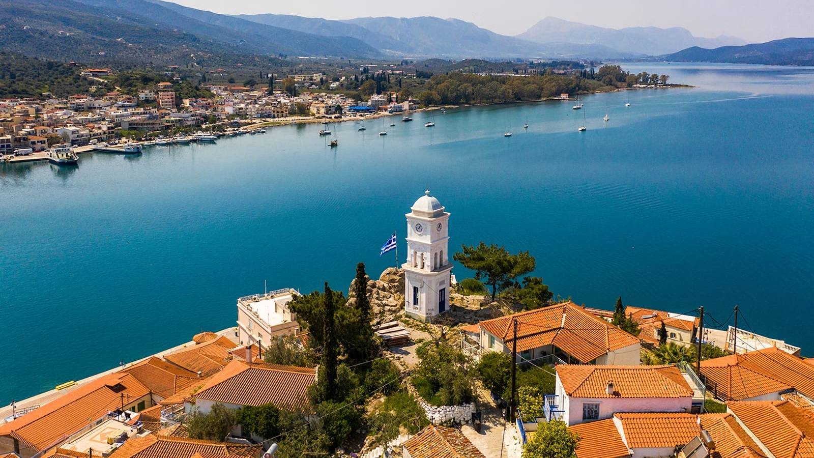 A charming clock tower stands sentinel over a picturesque Greek village nestled beside a tranquil turquoise bay.