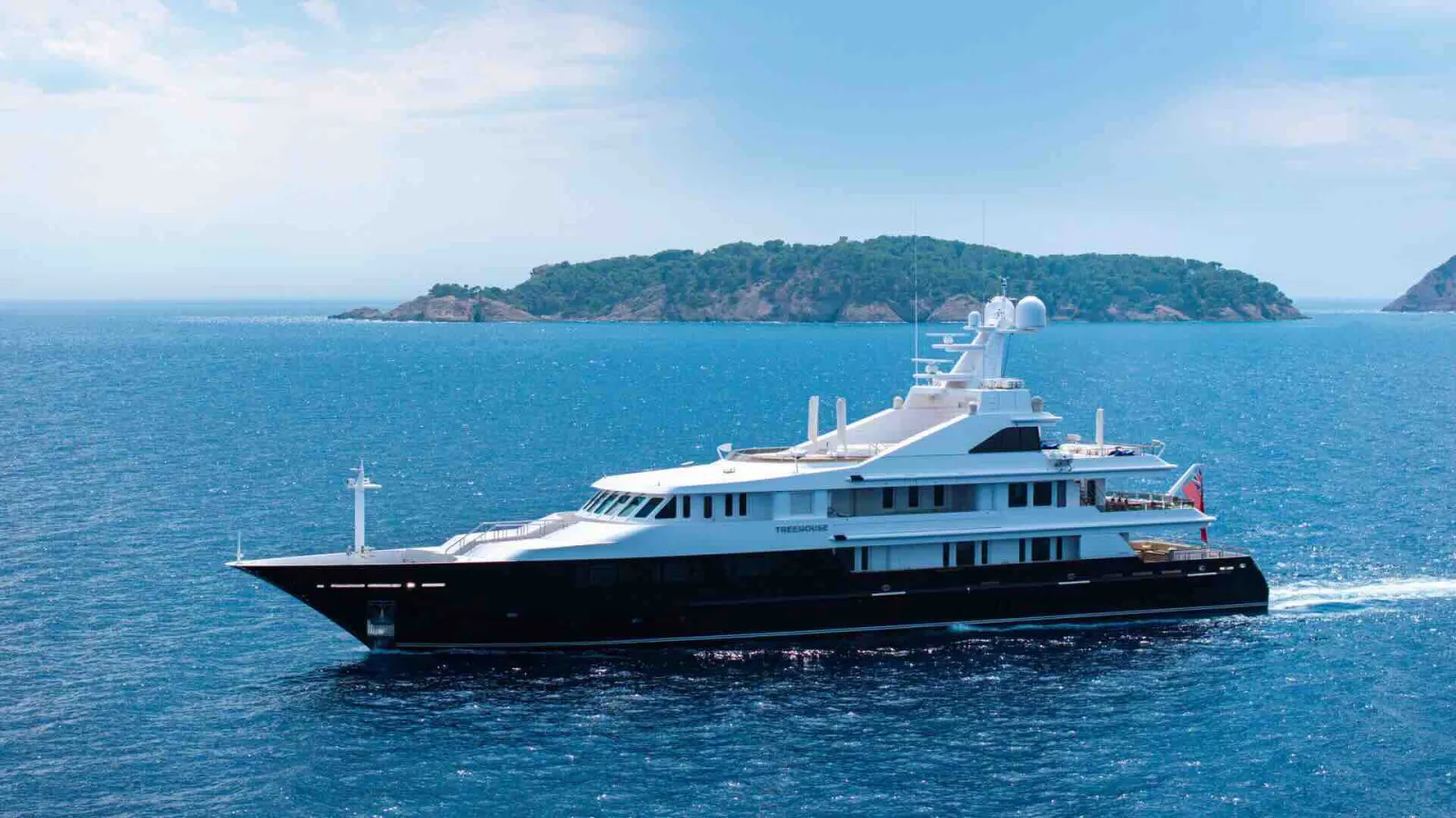 A sleek, dark-hulled yacht cruises on calm blue water with a lush, green island in the background under a hazy sky.