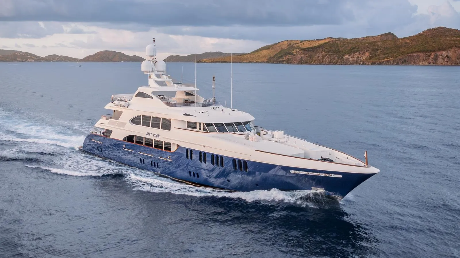 A large blue and white yacht cruises through calm water with a hilly, green coastline in the background.