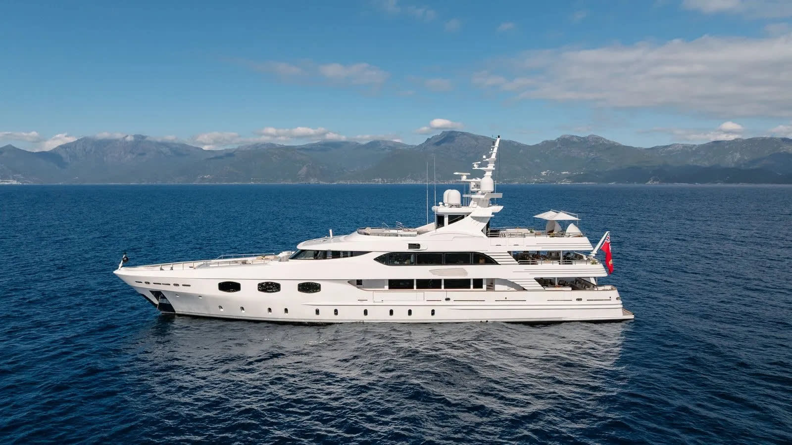 A large white yacht floats on calm blue water in the foreground, with a backdrop of distant, hazy mountains under a clear blue sky.