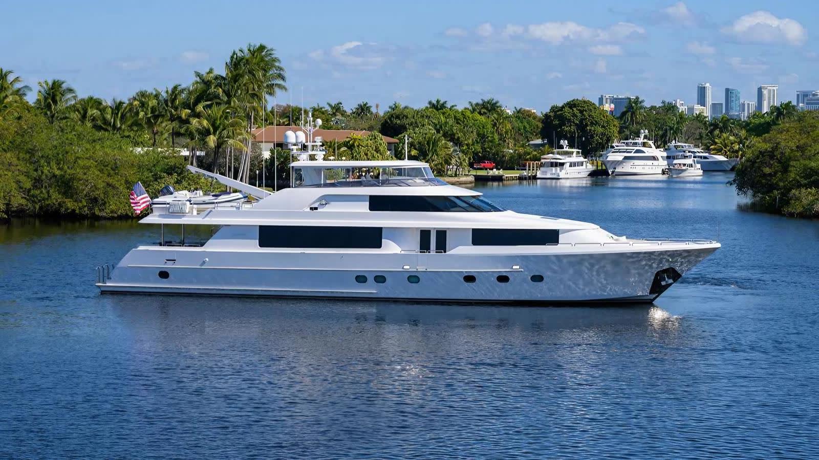 A sleek white yacht floats on a calm blue waterway, with lush tropical greenery and a distant city skyline under a bright sky.