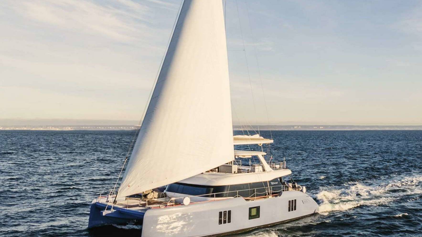A large white sailboat with a tall mast glides across the calm blue waters, with a scenic horizon visible in the background.