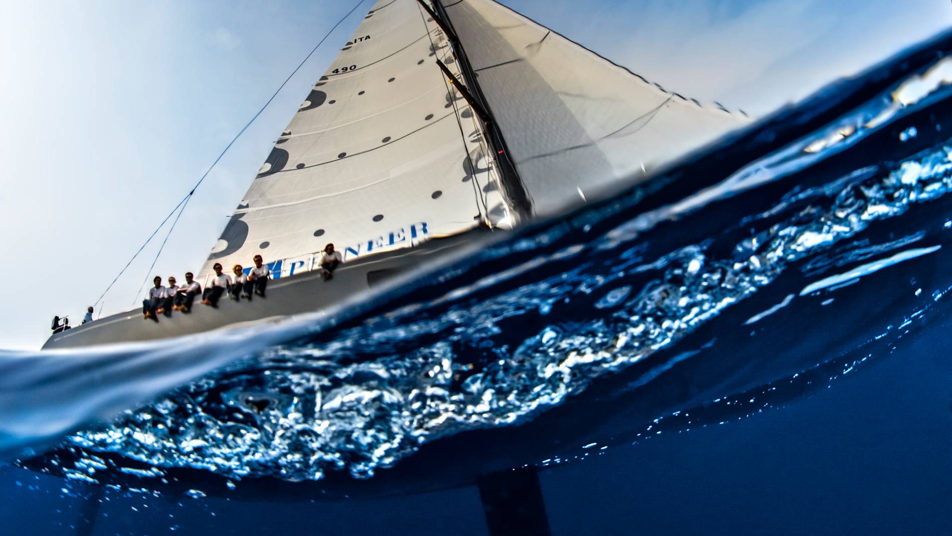 A sailboat's crew enjoys the ride as the hull cuts through the choppy blue ocean waves.