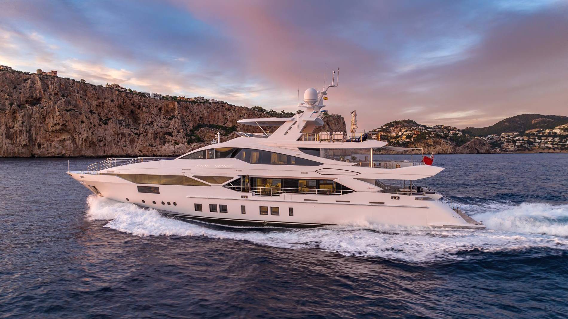 A luxurious white yacht cruises through the deep blue sea against a backdrop of rugged cliffs and a colorful sunset sky.