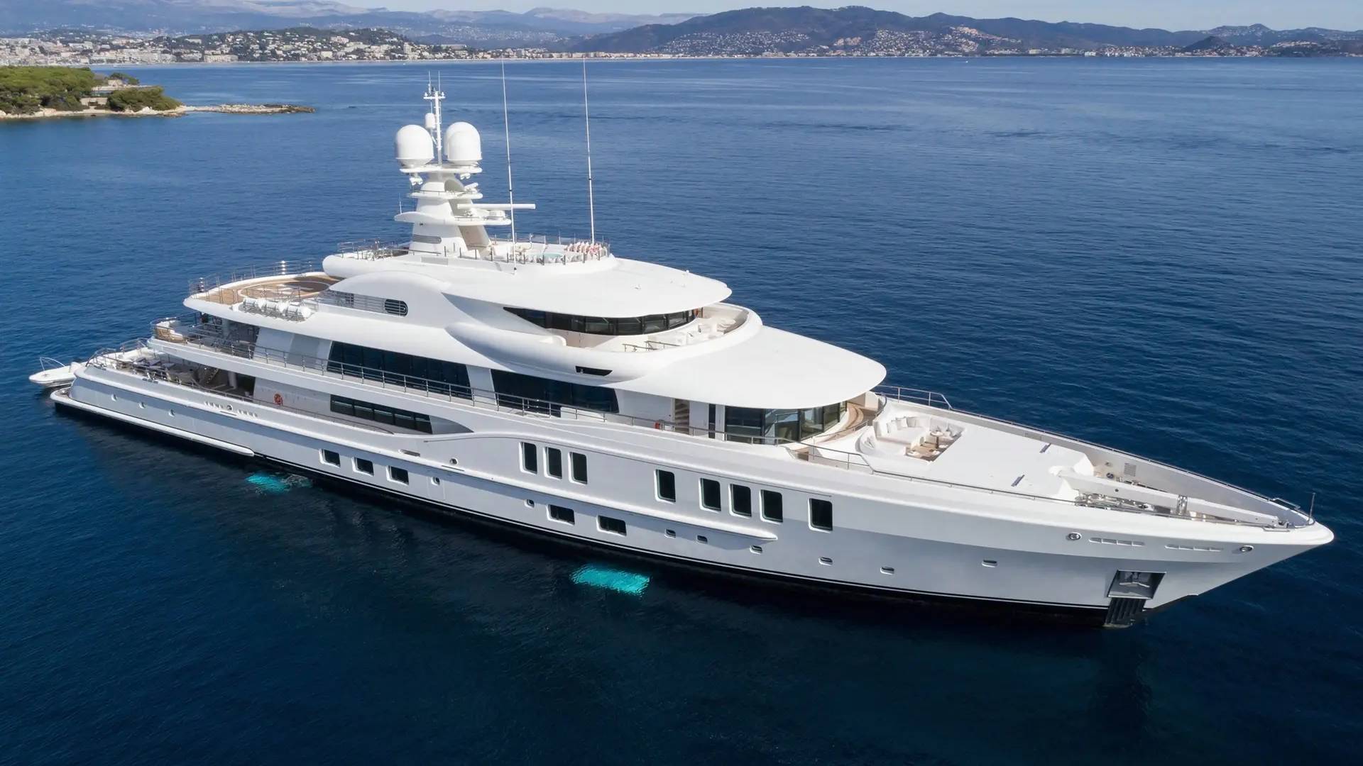 A luxurious white superyacht glides through the deep blue ocean, with a picturesque coastline in the background.