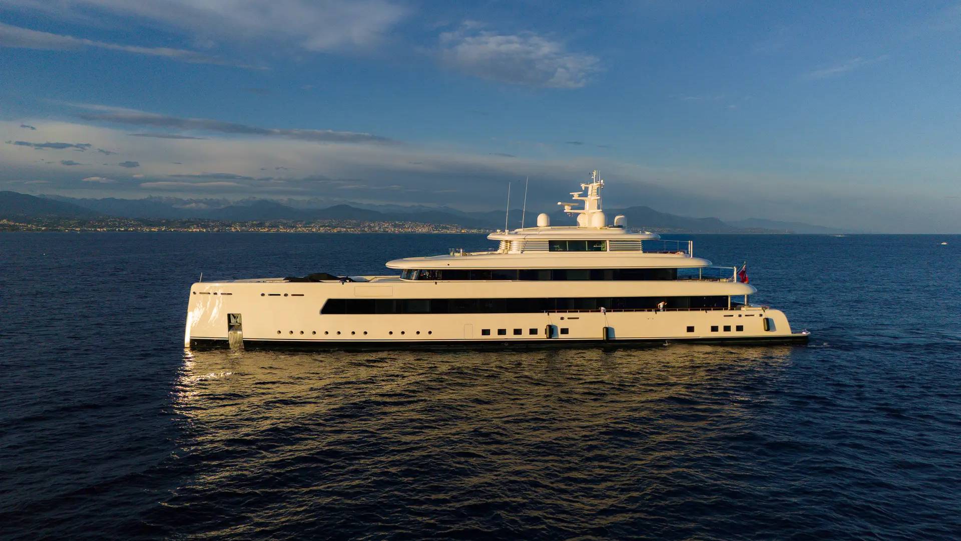 A luxurious white yacht glides serenely on the deep blue sea under a partly cloudy sky with distant mountains.