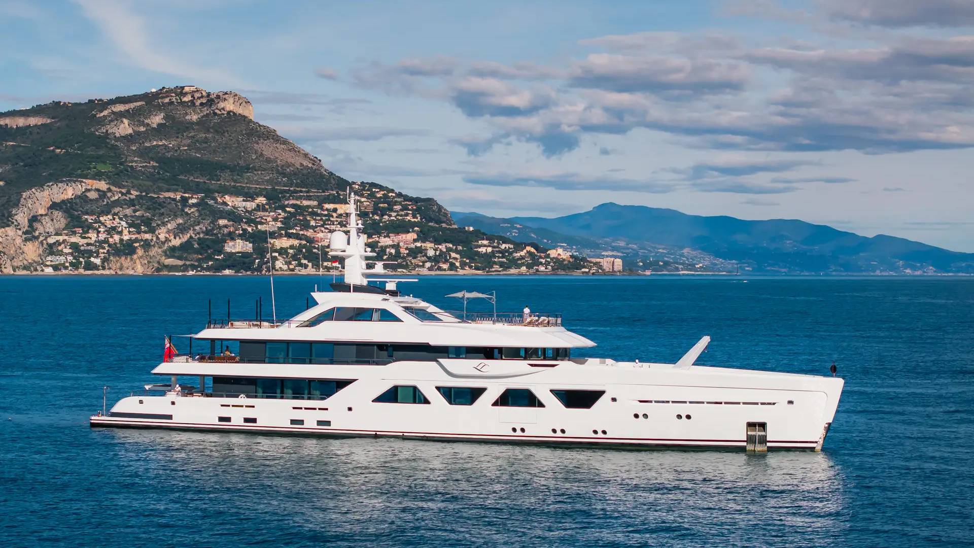 A luxurious white yacht cruises serenely on the azure waters off a picturesque coastal town nestled against rolling hills.