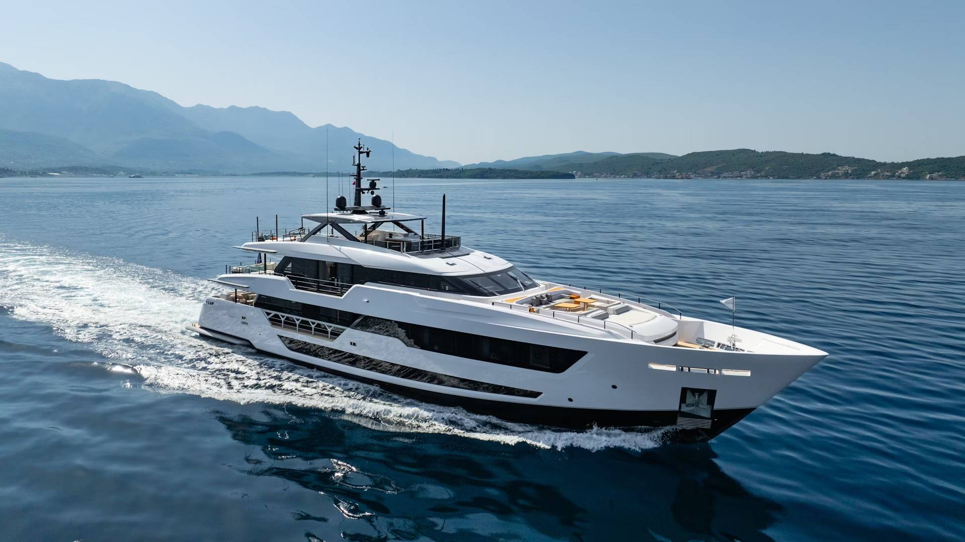 A sleek white yacht cruises through calm blue waters with distant mountains under a clear sky.