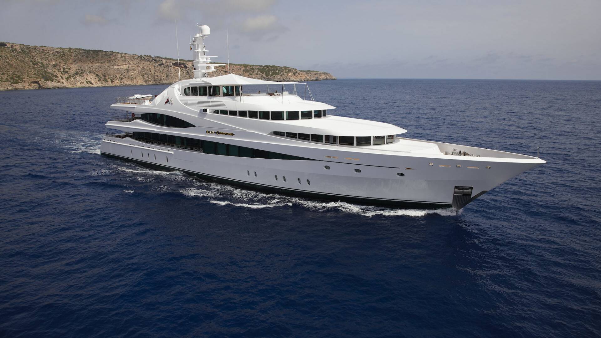 A luxurious white yacht glides through the deep blue ocean near a rocky coastline under a cloudy sky.