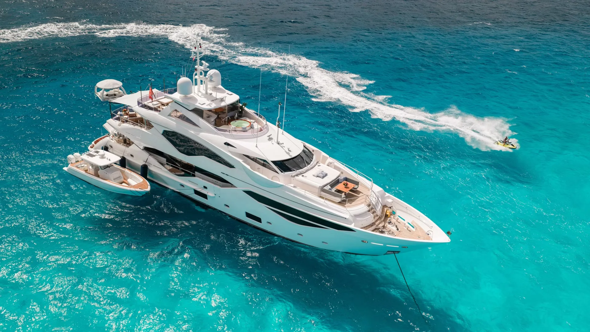 A large, luxurious yacht cruising through the crystal-clear, turquoise waters of what appears to be a tropical or subtropical location, with a wake of white foam trailing behind it.