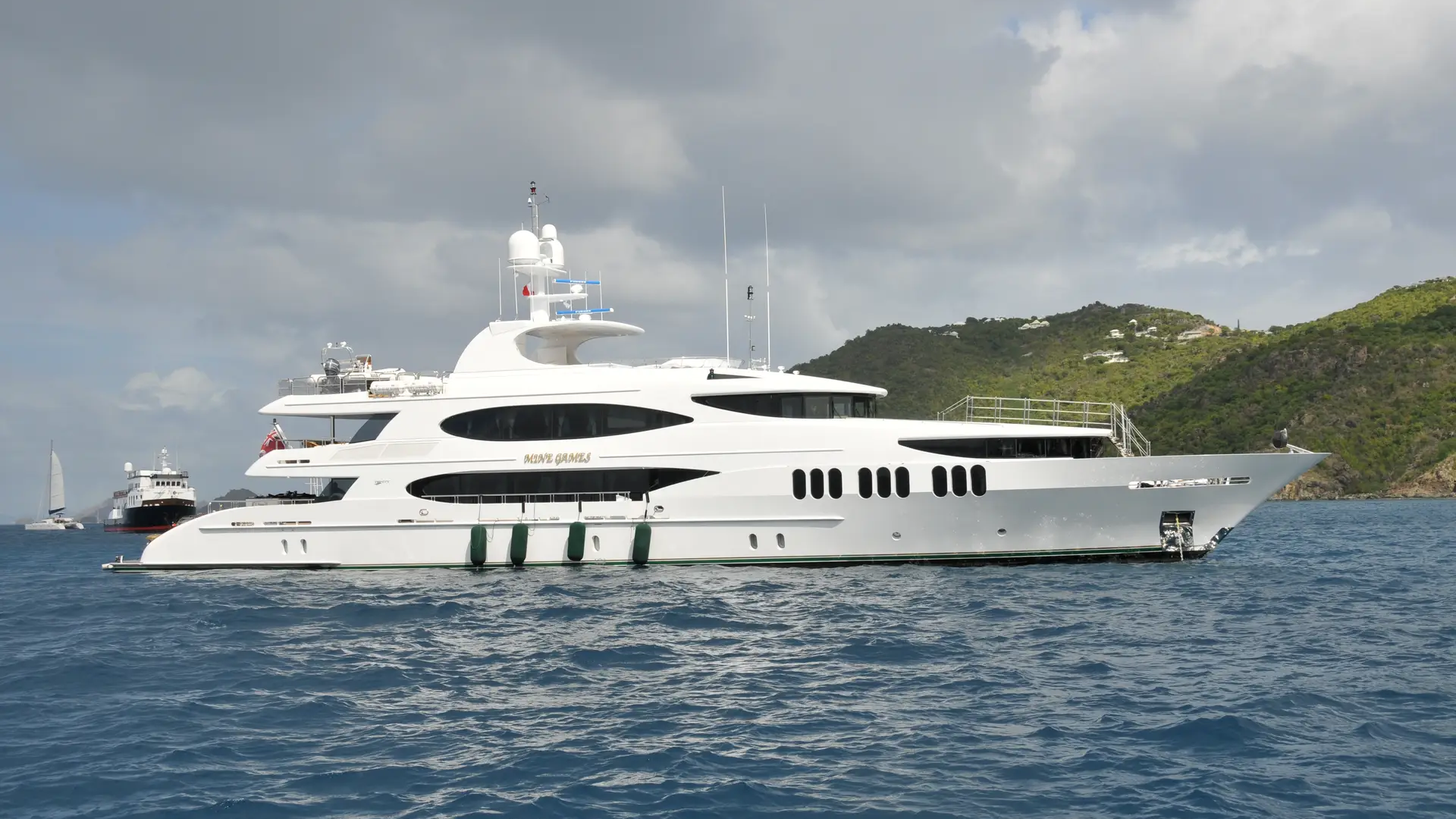 A large white yacht with green trim sails on blue water in the foreground, while a lush green island with scattered buildings and a cloudy sky form the background.