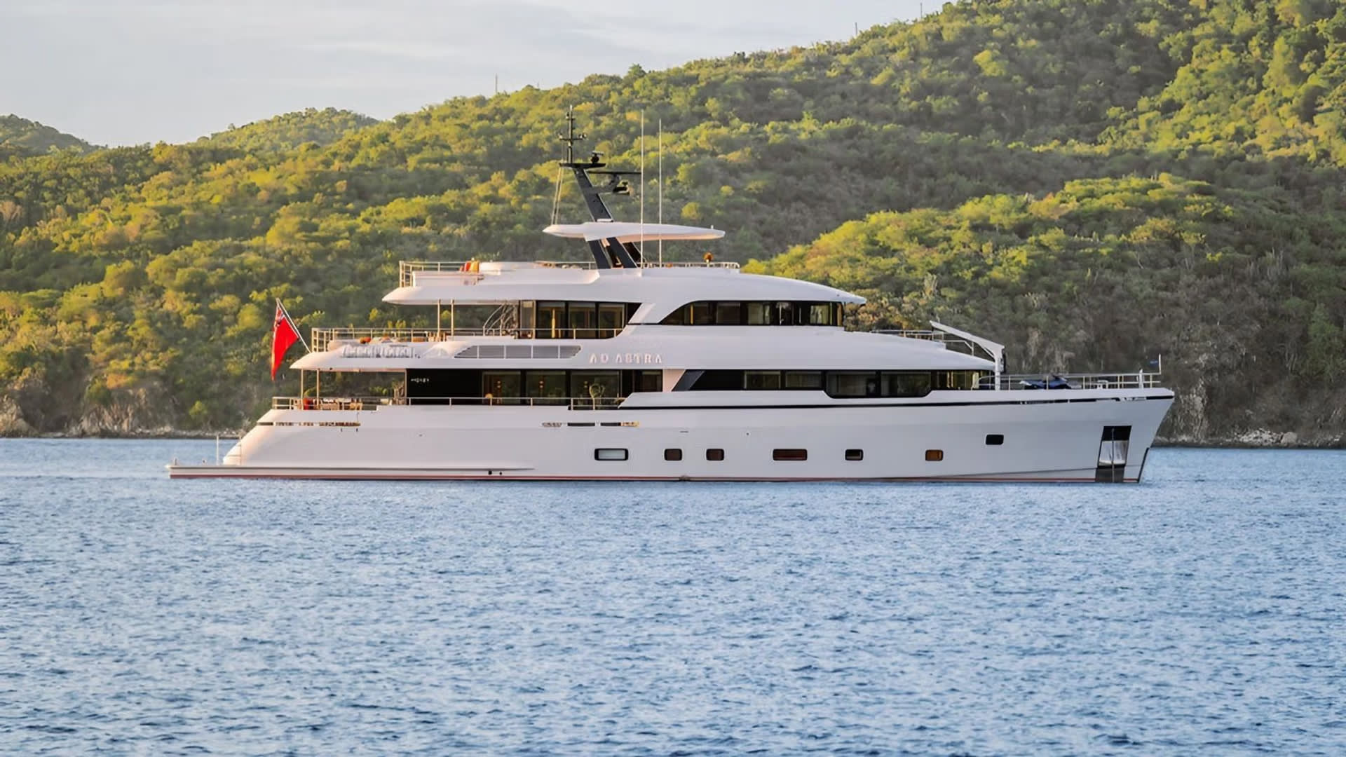 A large white yacht sails on calm blue water in the foreground, with lush green hills rising in the background.