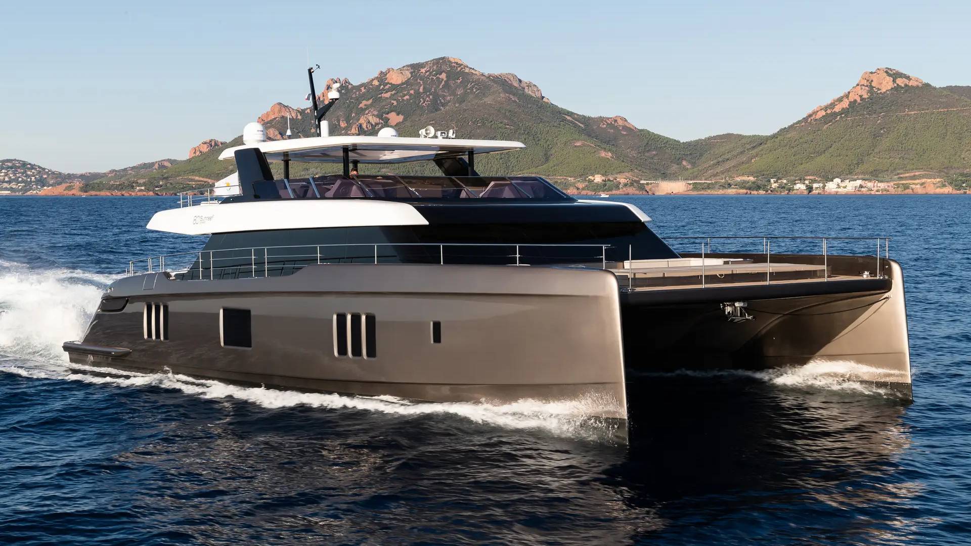 A sleek, modern catamaran yacht cruises through calm blue water with a rugged, mountainous coastline in the background.