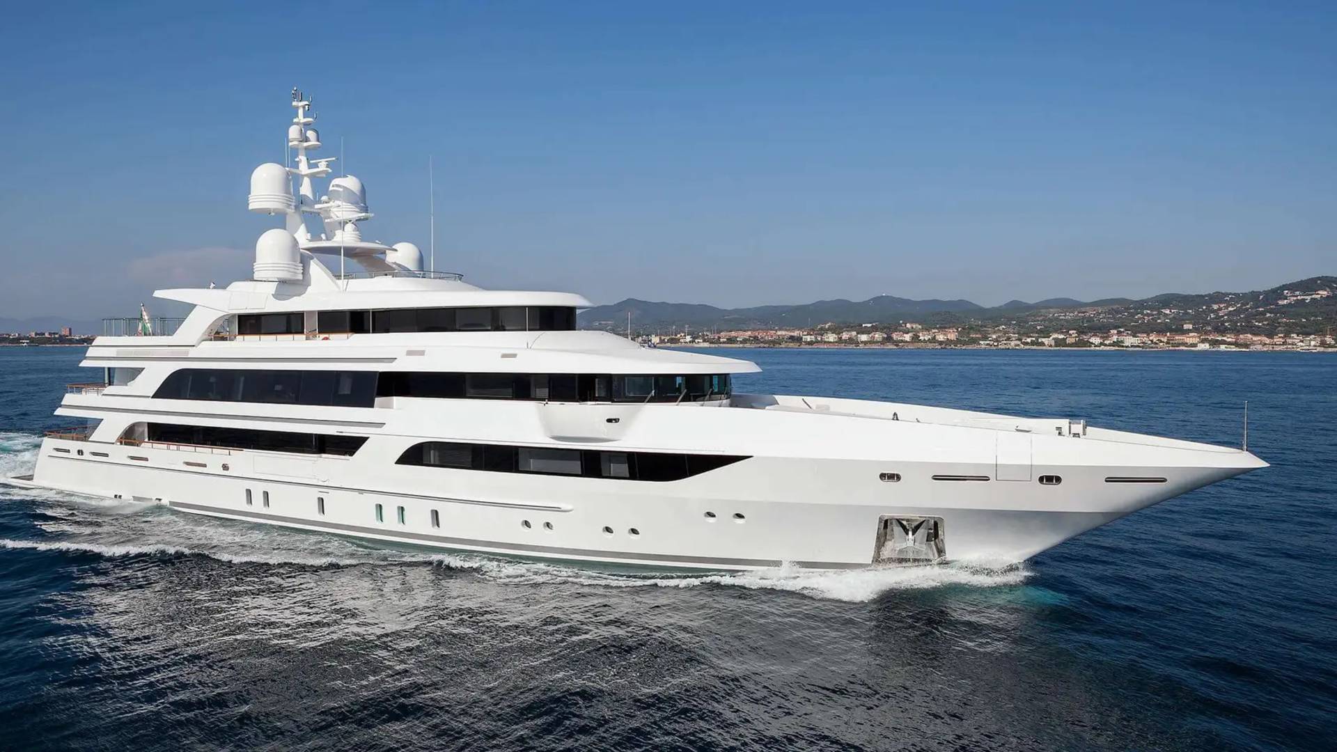 A large, white luxury yacht cruises through deep blue water with a distant coastline and mountains under a clear sky.
