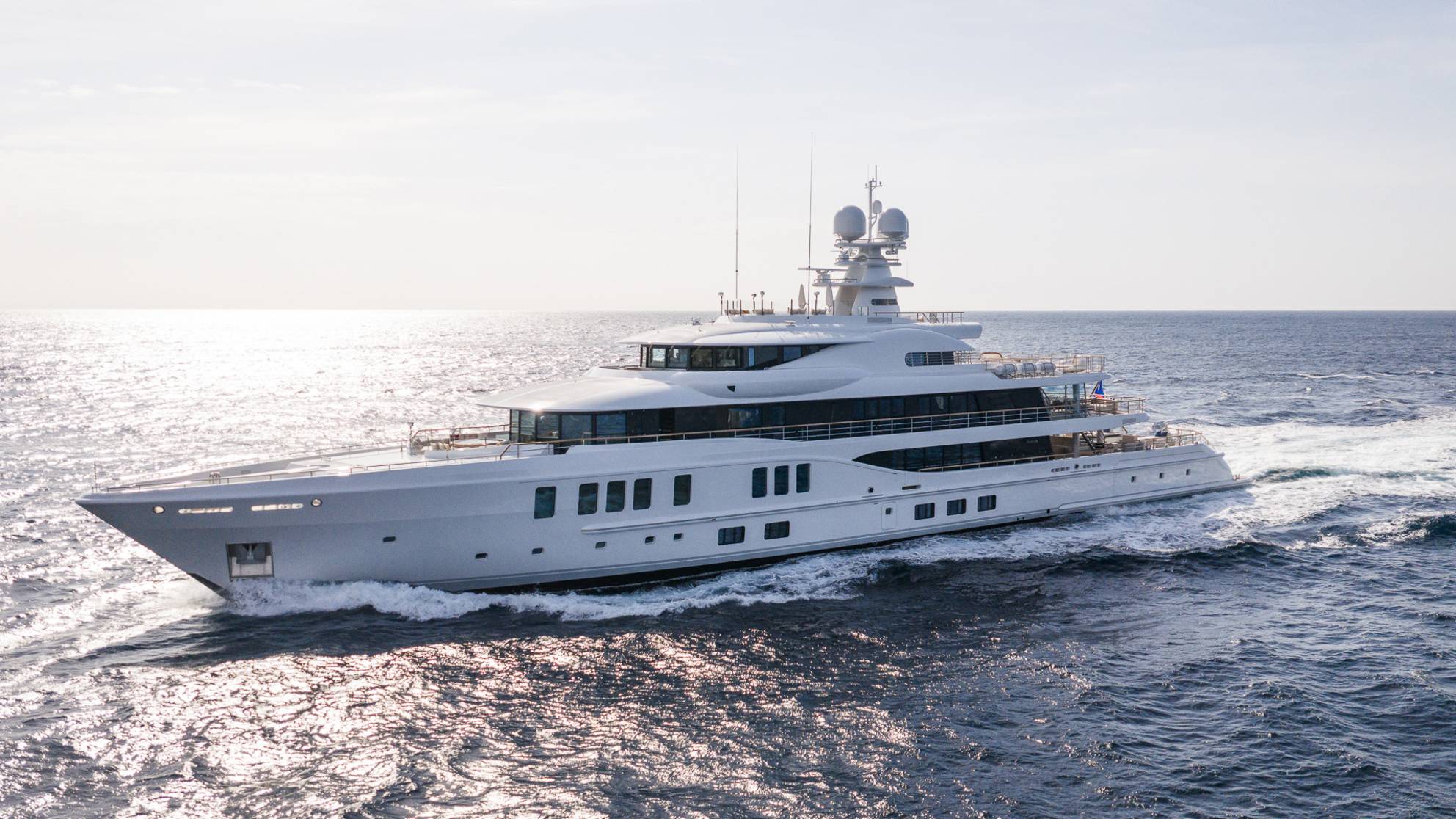 A large white yacht cruises through calm, sun-dappled ocean waters under a bright sky.