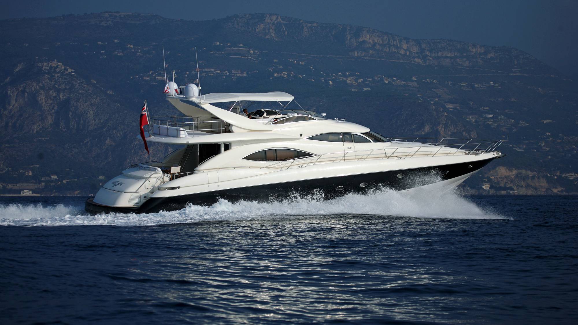 A sleek white yacht cuts through the deep blue water, leaving a foamy wake against a backdrop of rugged, tree-covered hills.