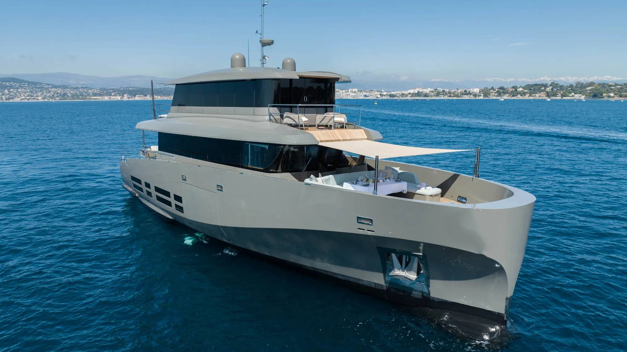 A sleek, modern gray yacht with an open deck area is anchored in calm blue water, with a distant coastline and clear sky in the background.