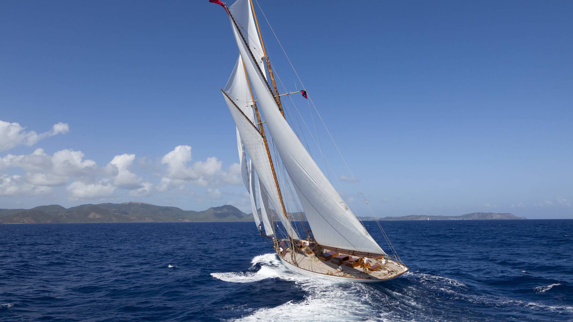 A classic sailboat with white sails cuts through the deep blue ocean, leaving a foamy wake, with distant, hazy islands under a clear blue sky.