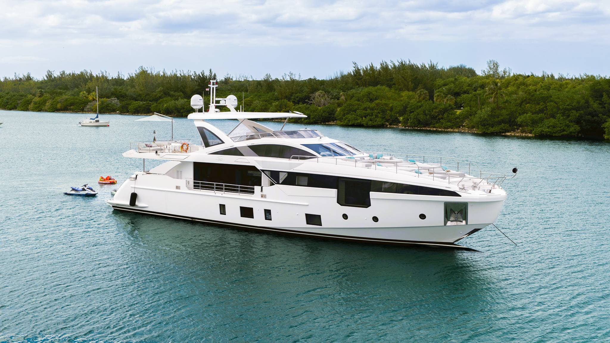 A sleek white yacht floats on calm blue water in the foreground, with lush green trees and a cloudy sky forming the background.