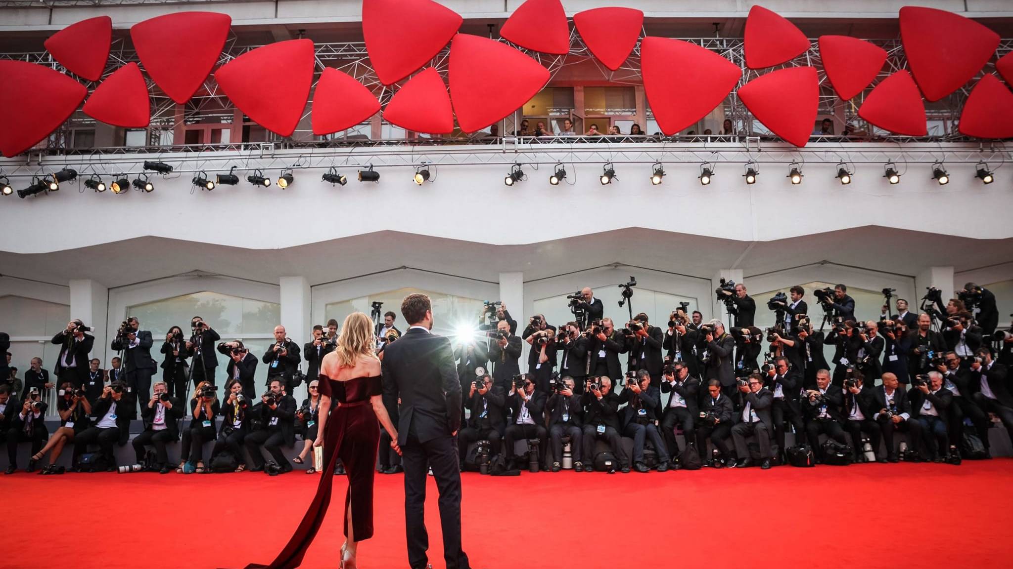 A couple in formal attire walks hand-in-hand on a red carpet, surrounded by a throng of photographers, with a backdrop of red abstract sculptures and tiered lighting.