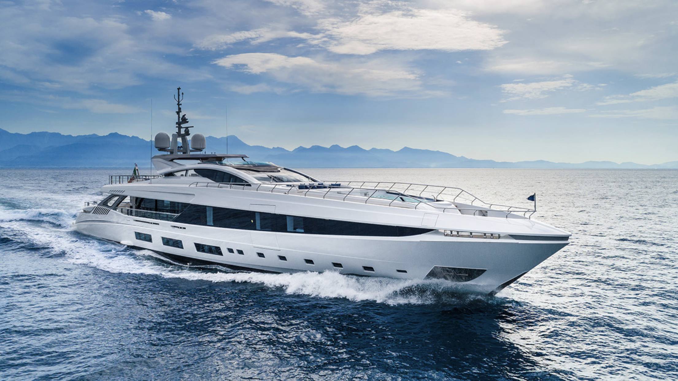 A sleek white yacht cuts through the deep blue ocean waters with mountains in the background.