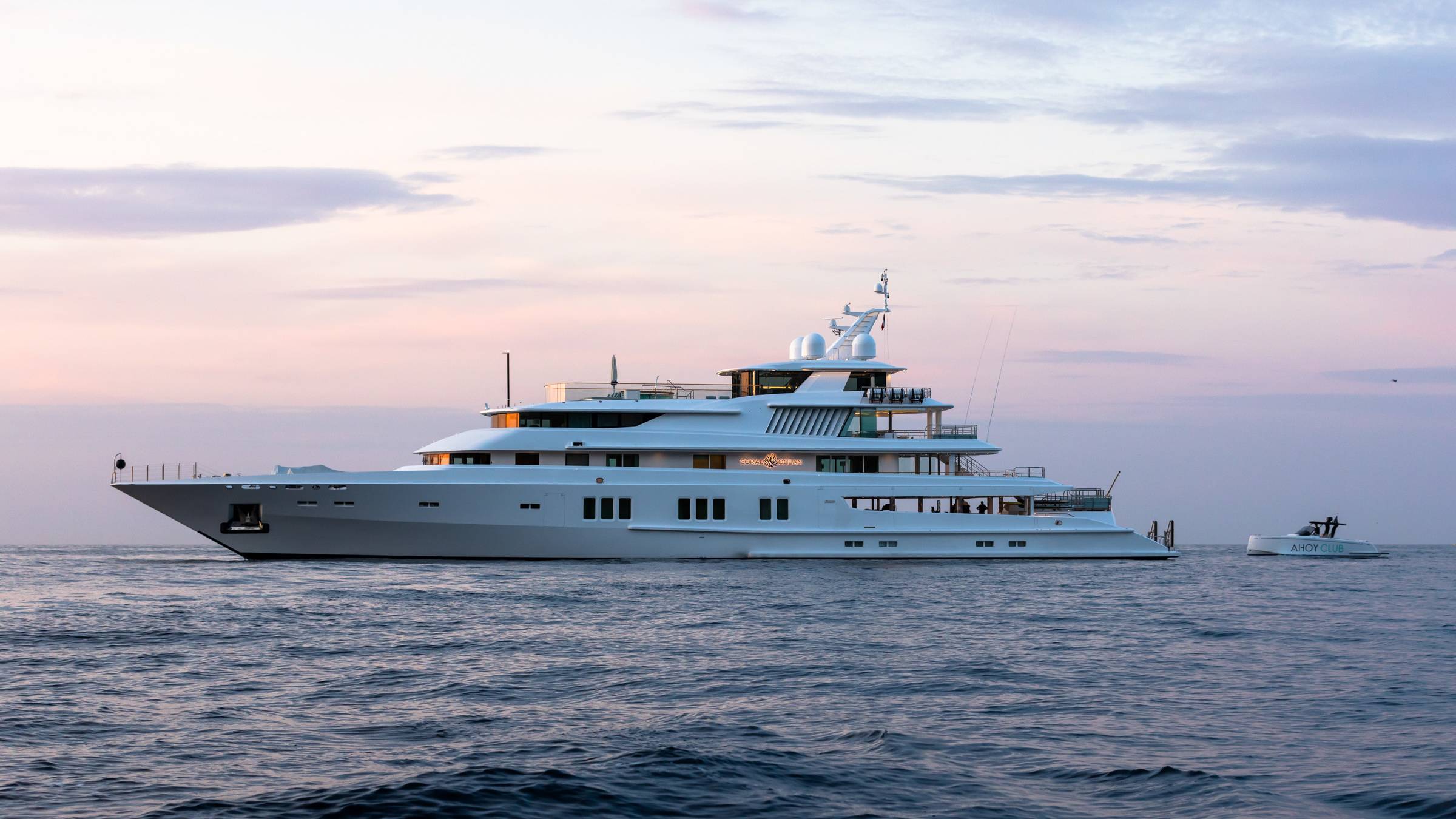 A luxurious white superyacht named Coral Ocean glides across calm waters under a soft, pastel sky.