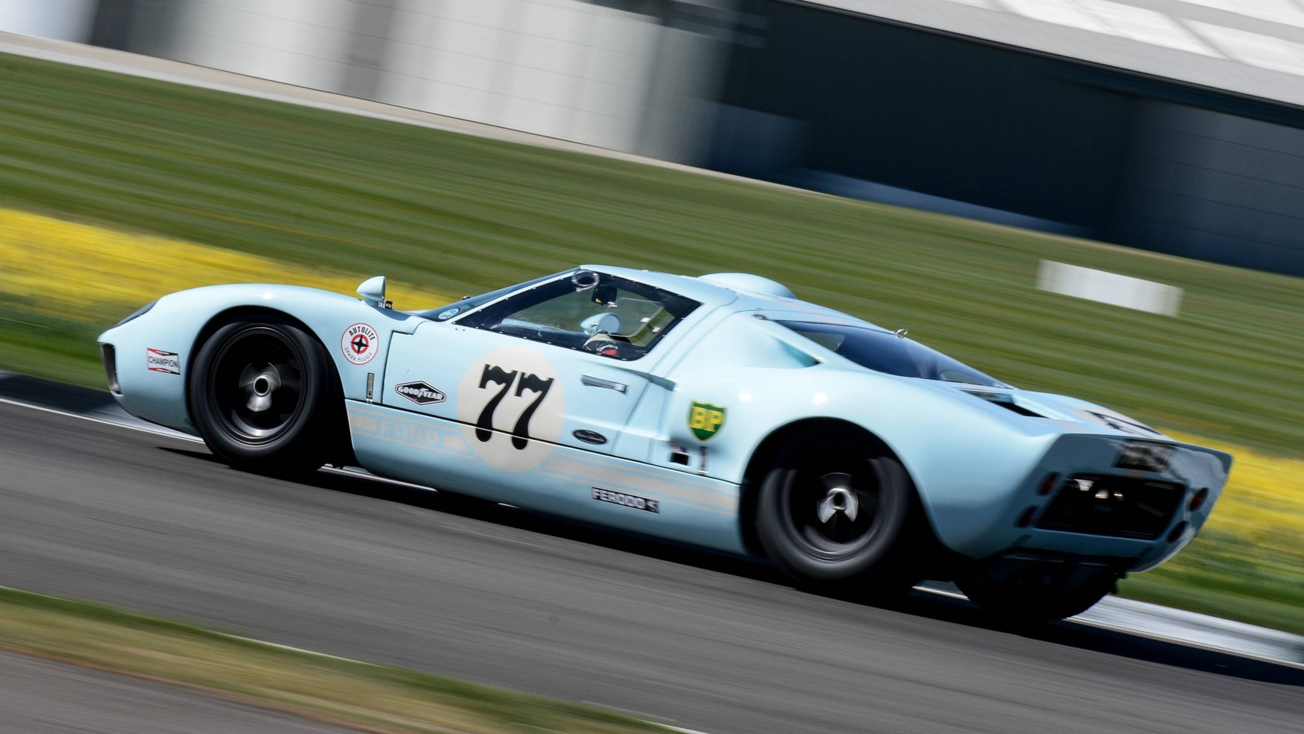 A light blue Ford GT40 races down the track, its number 77 prominently displayed.