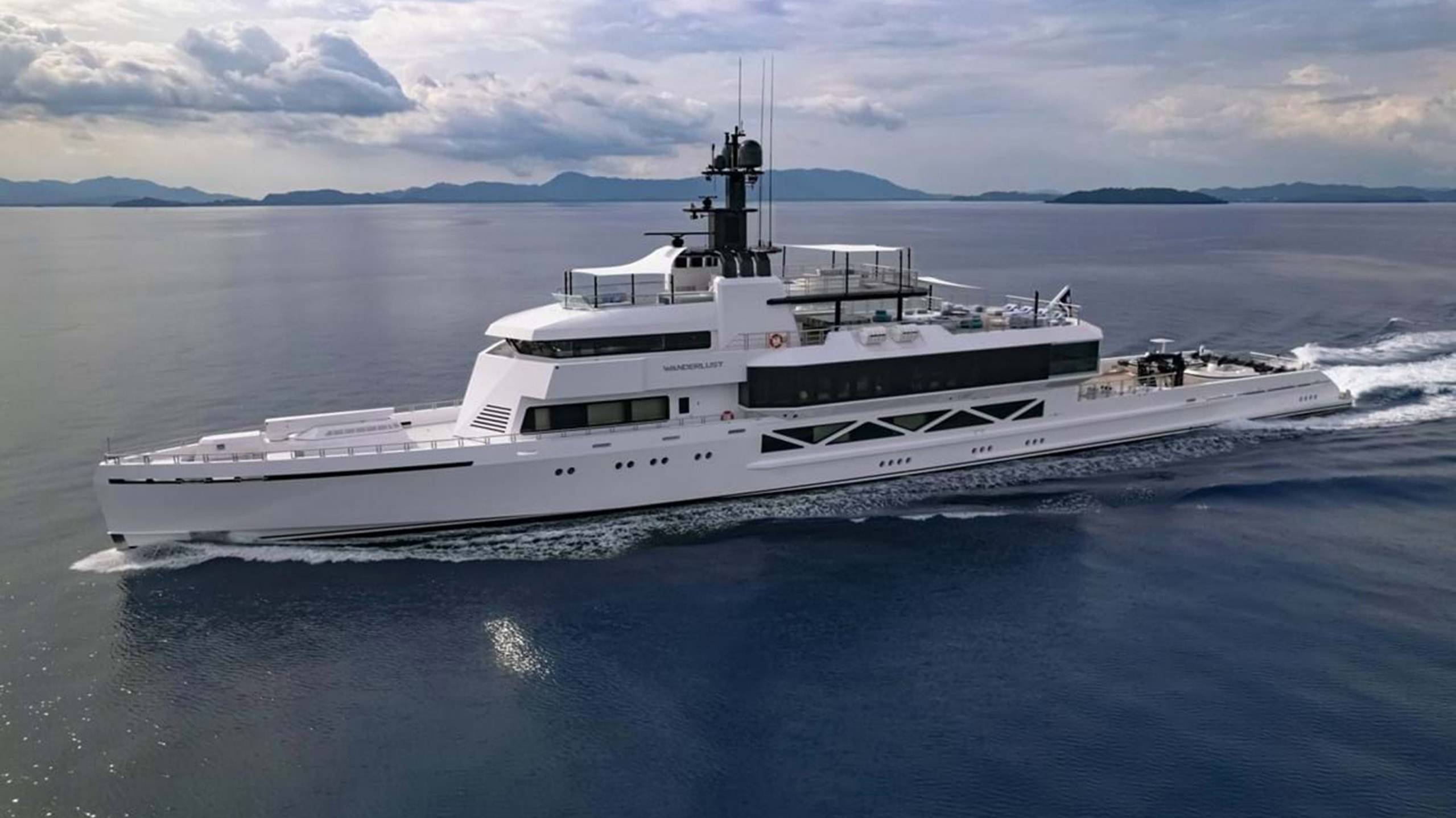 The sleek white yacht Wanderlust glides through calm blue waters beneath a dramatic cloudy sky.