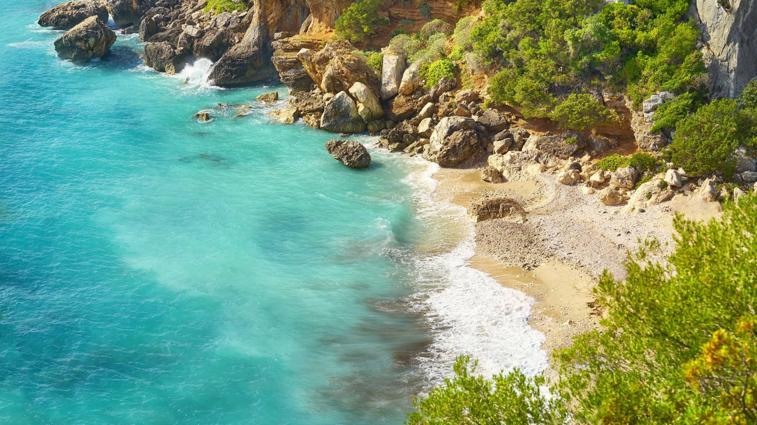 Turquoise waves crash against the rocky shore of a secluded Mediterranean cove, fringed by lush green foliage.