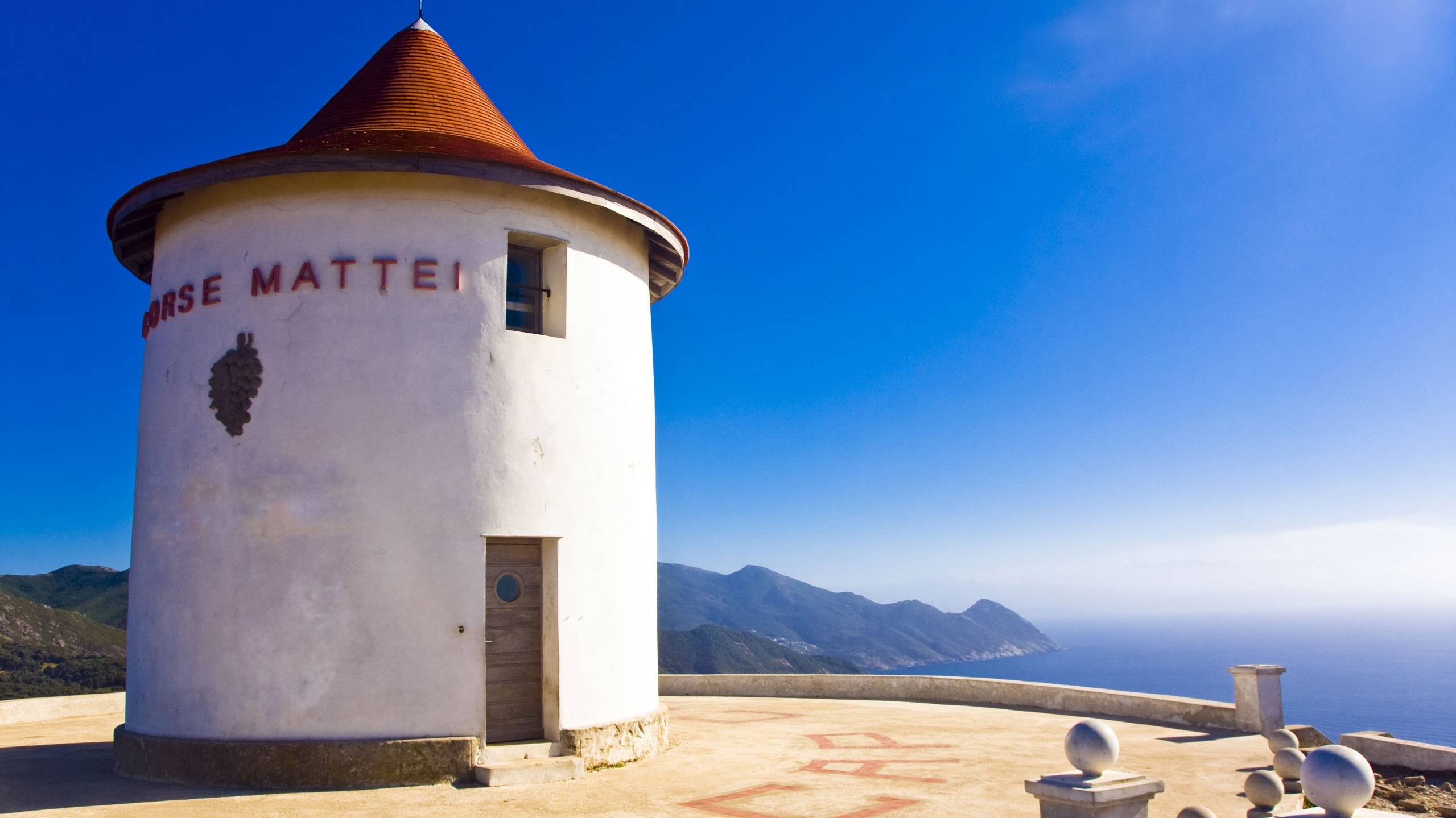 A white windmill with a red roof stands on a clifftop overlooking the sparkling blue sea and distant mountains.