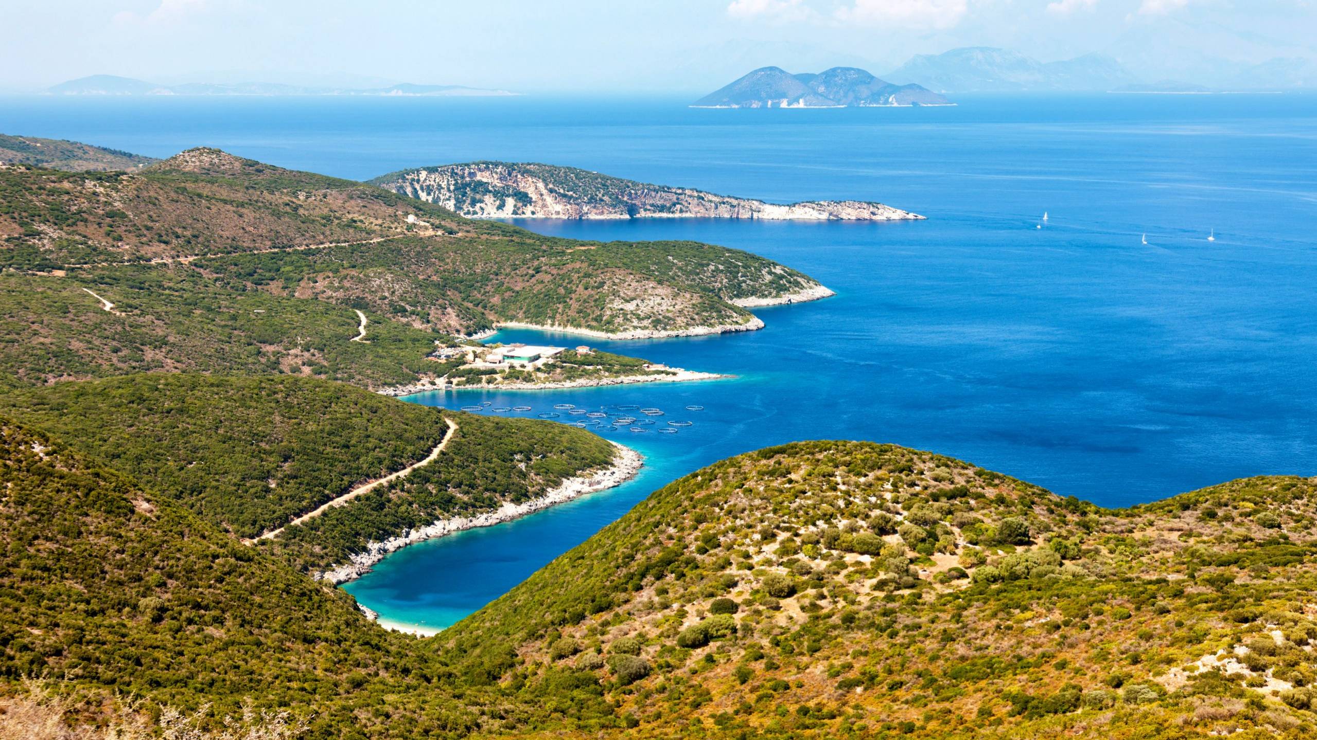 Lush green hills cascade down to a vibrant blue bay dotted with fish farms and distant sailboats.