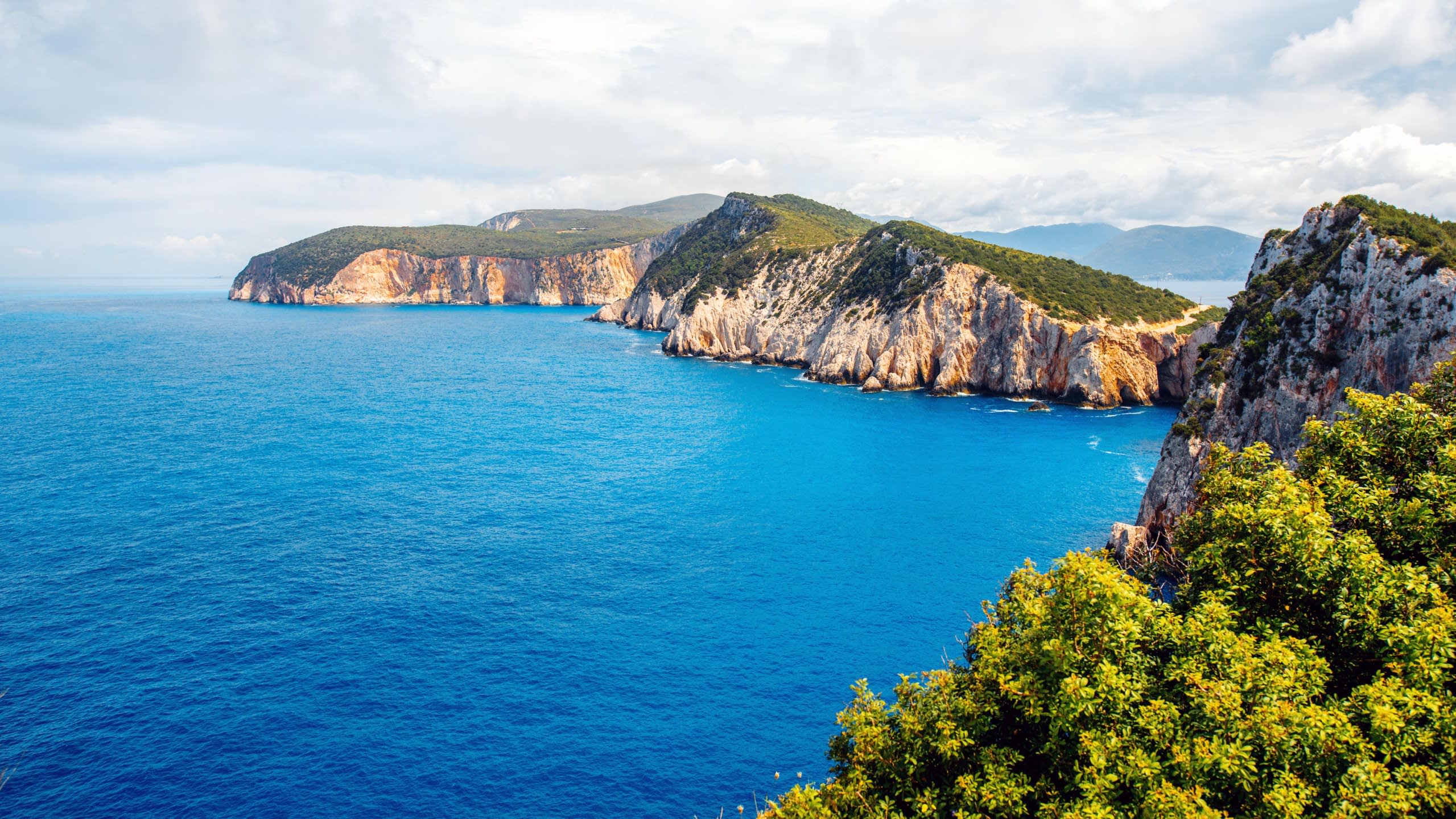 Dramatic cliffs plunge into the vibrant blue waters of the Ionian Sea under a cloudy sky.