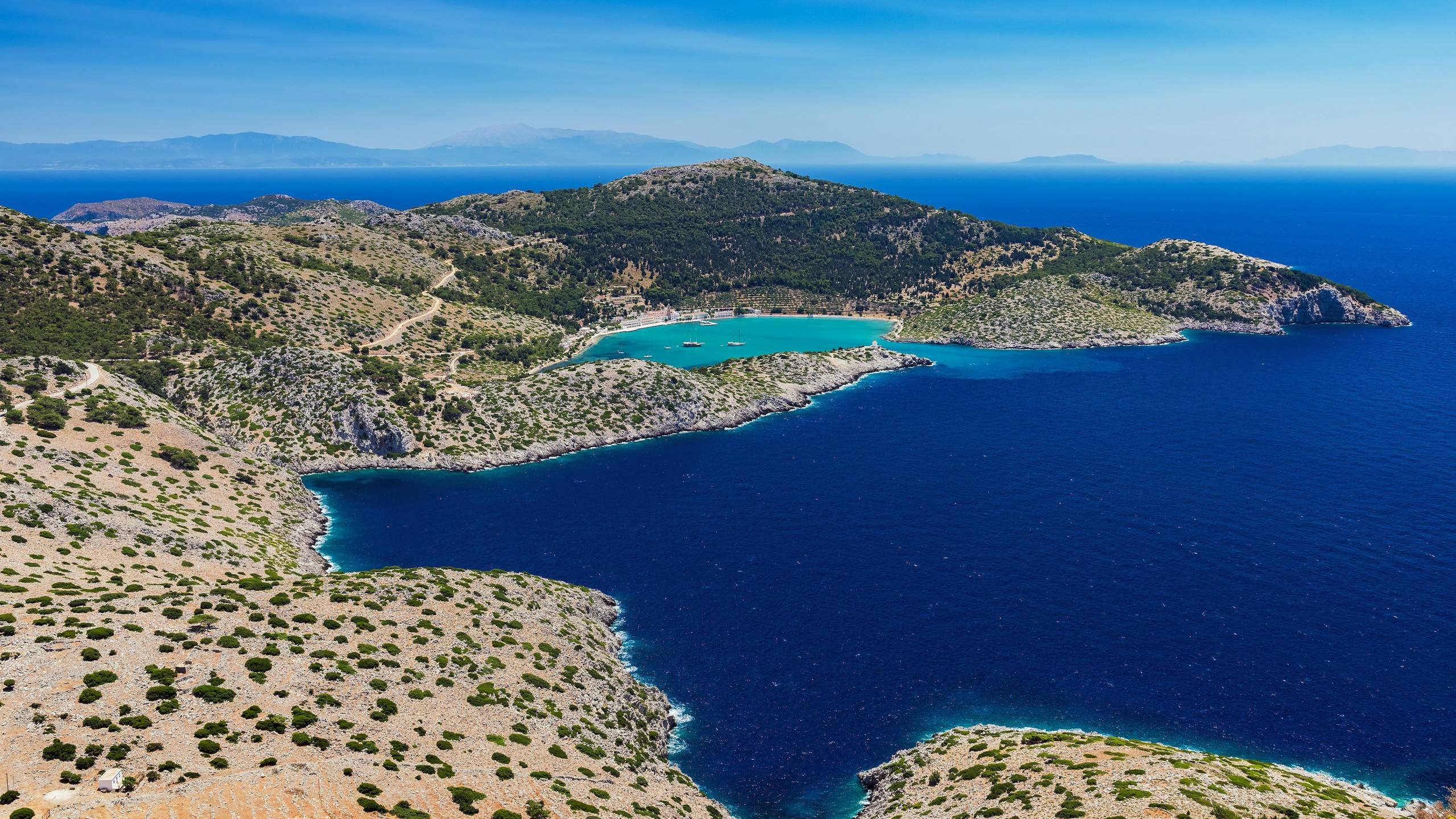 A stunning aerial view captures a turquoise bay nestled between rugged, green-covered hills under a clear blue sky.