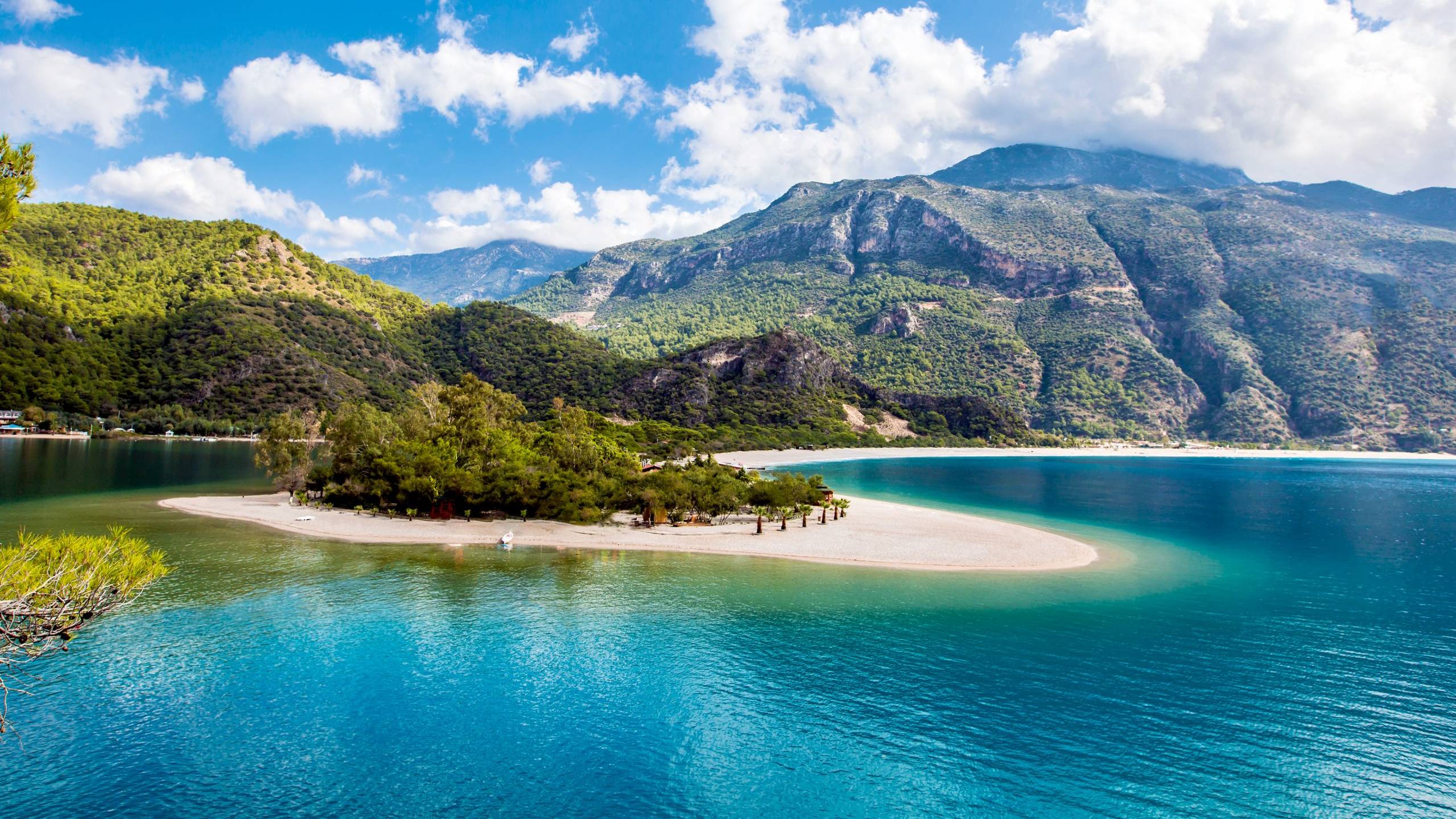 A crescent of white sand juts into the vibrant turquoise waters of a serene bay, backed by lush green mountains under a bright blue sky.