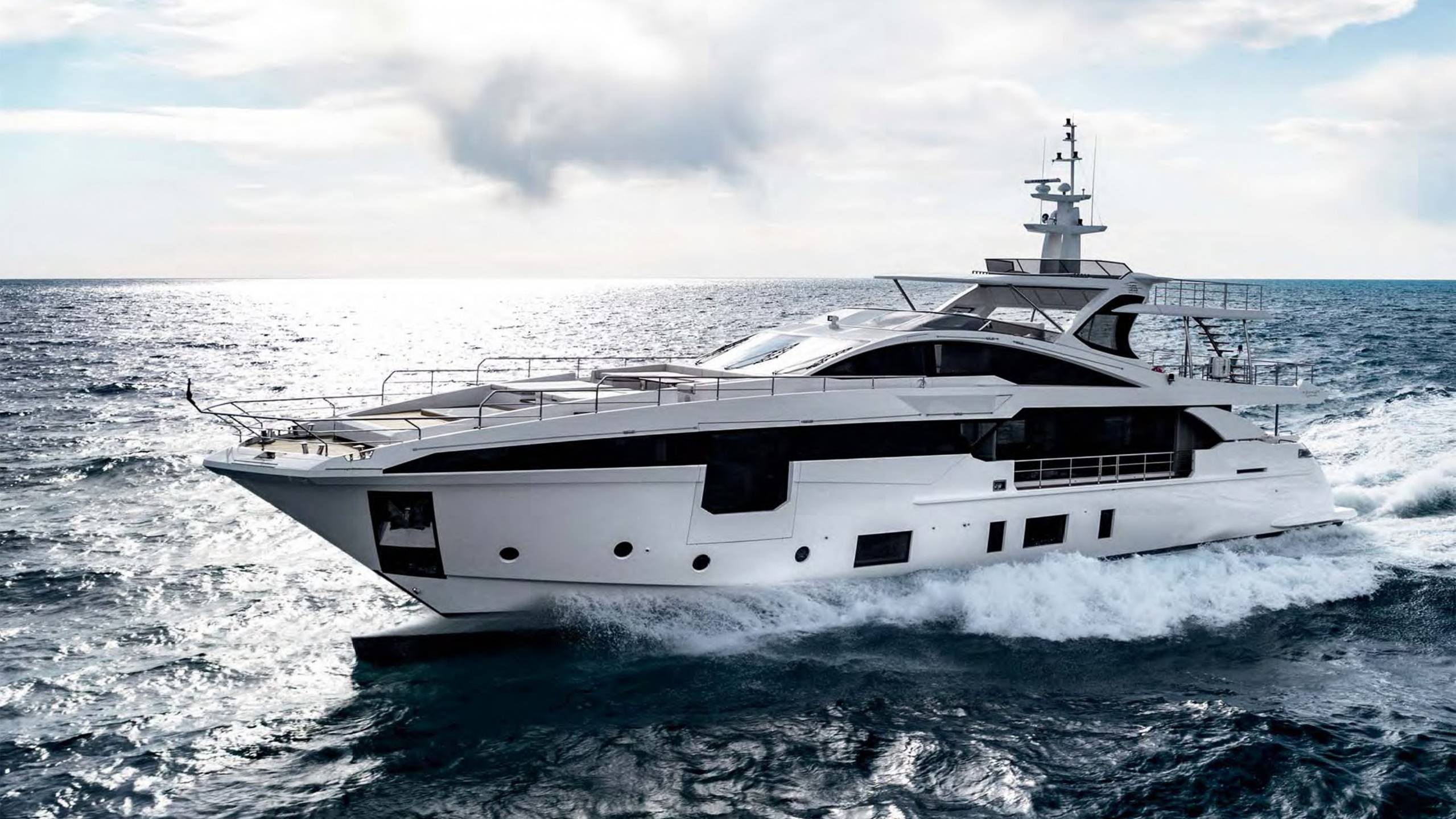 A sleek white yacht cuts through the choppy blue waters under a bright, cloudy sky.