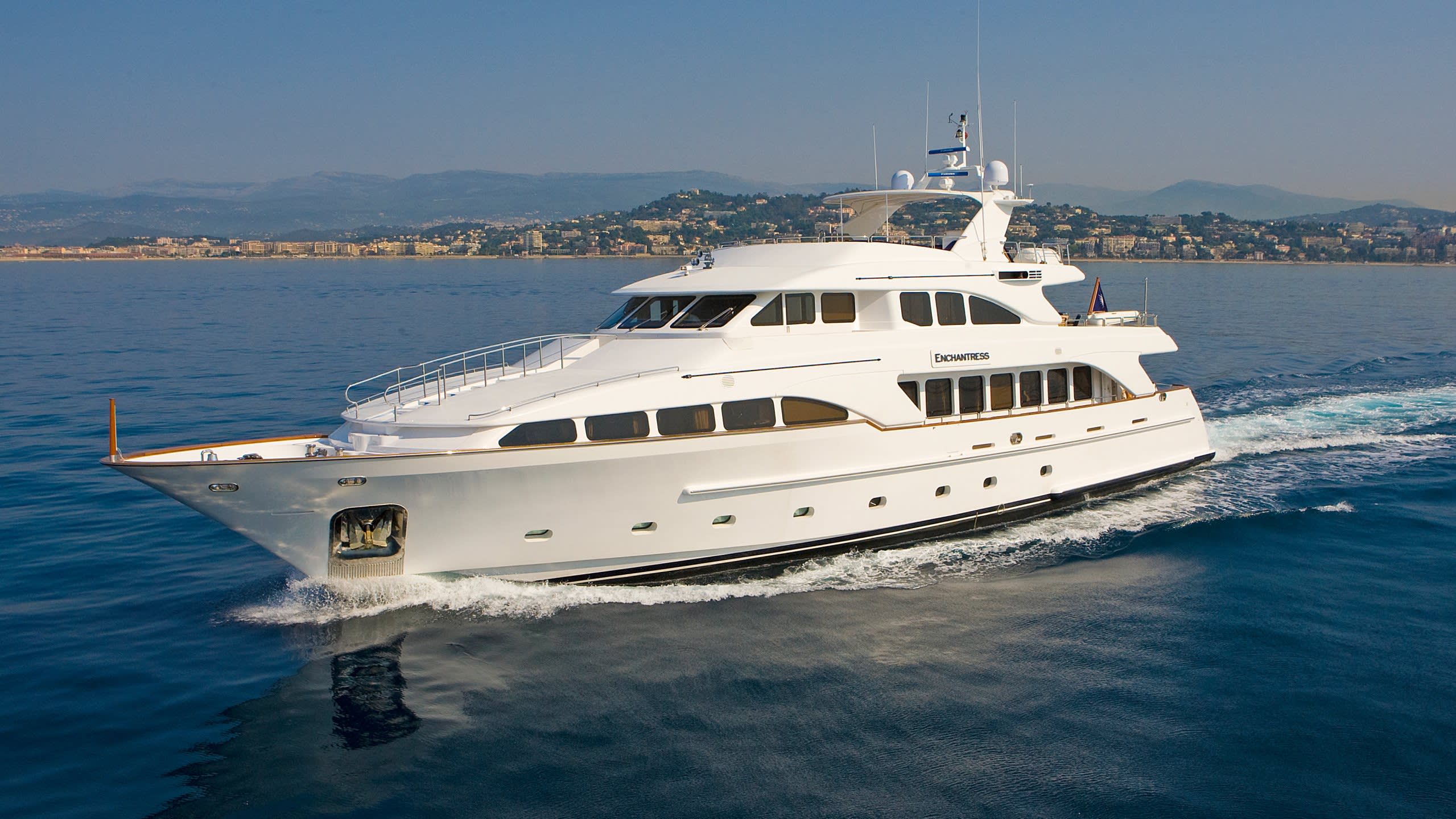 The luxurious yacht Enchantress glides through the calm blue waters with a picturesque coastal town in the background.
