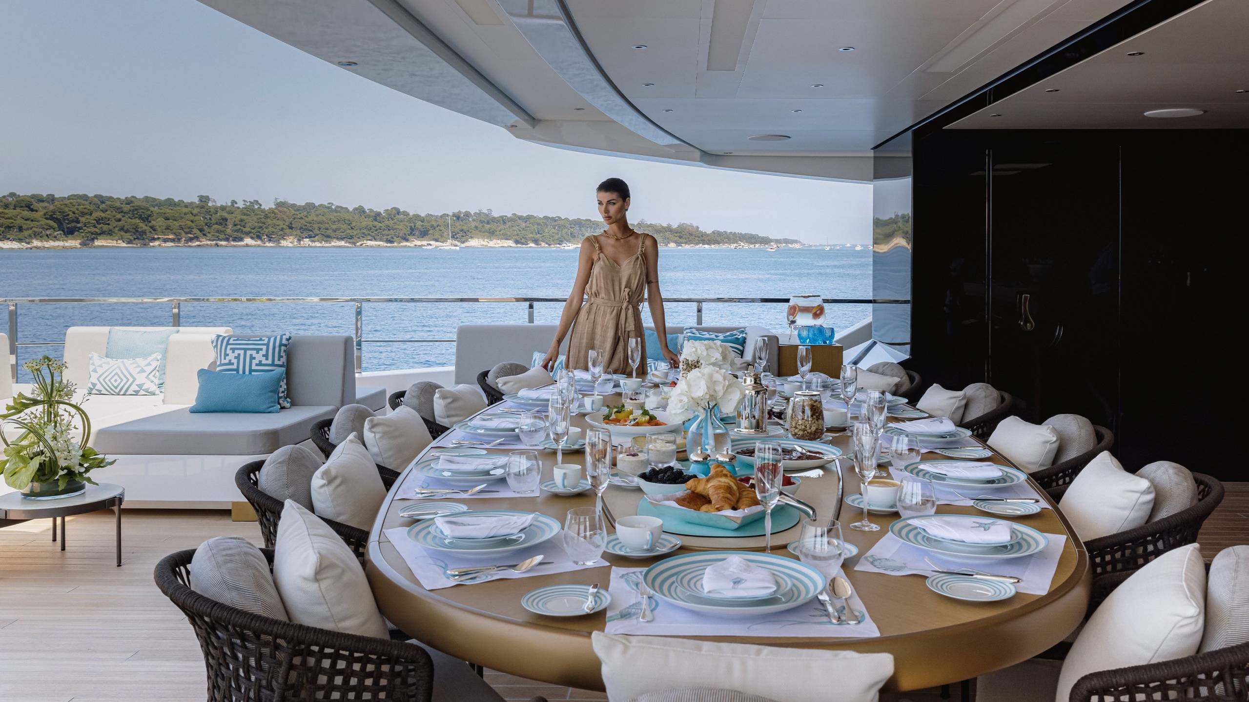 A woman in a beige dress stands by a beautifully set dining table on a yacht overlooking a serene blue ocean.