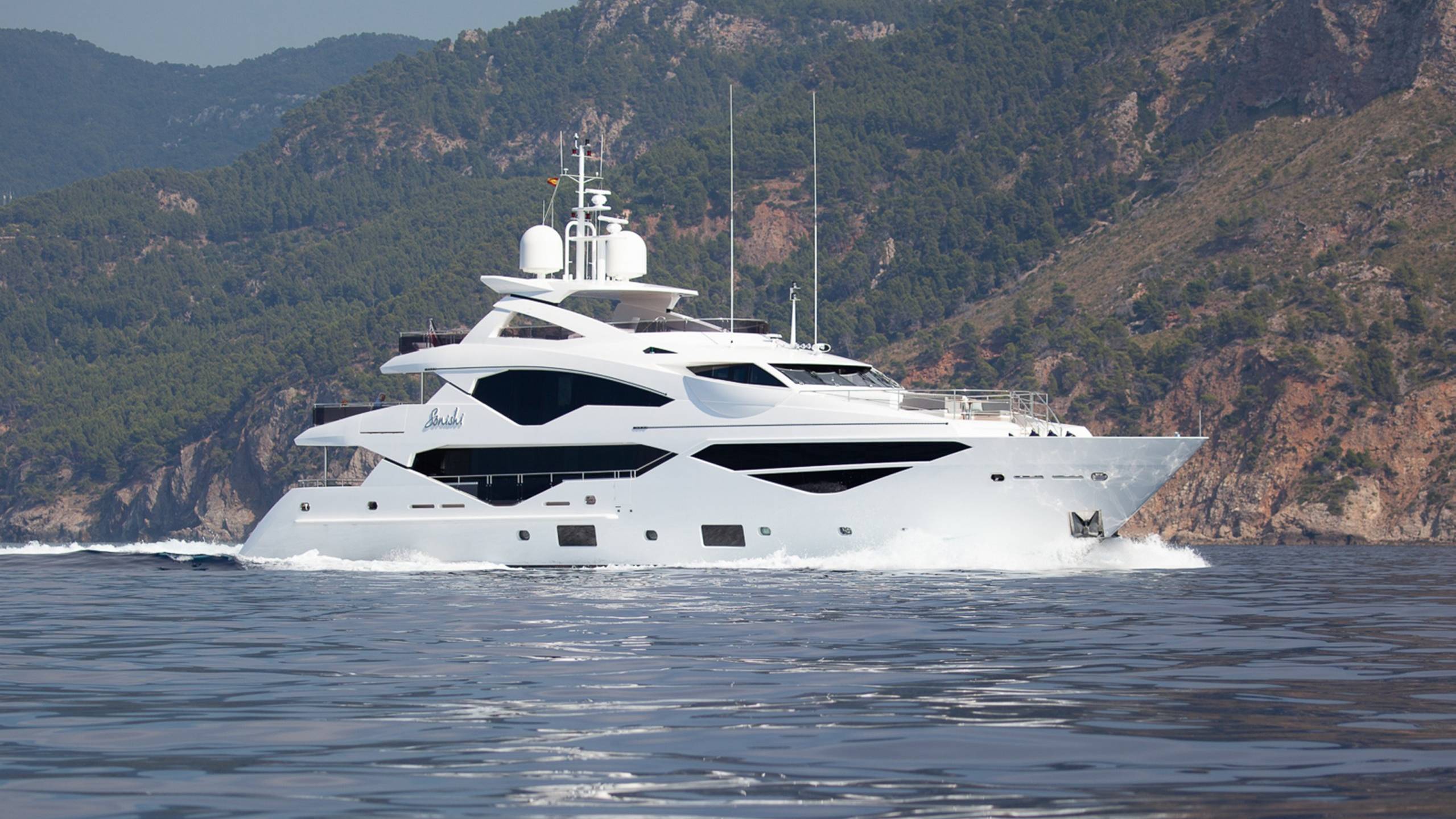 A sleek white yacht glides through calm waters with a lush, green mountainside as its backdrop.