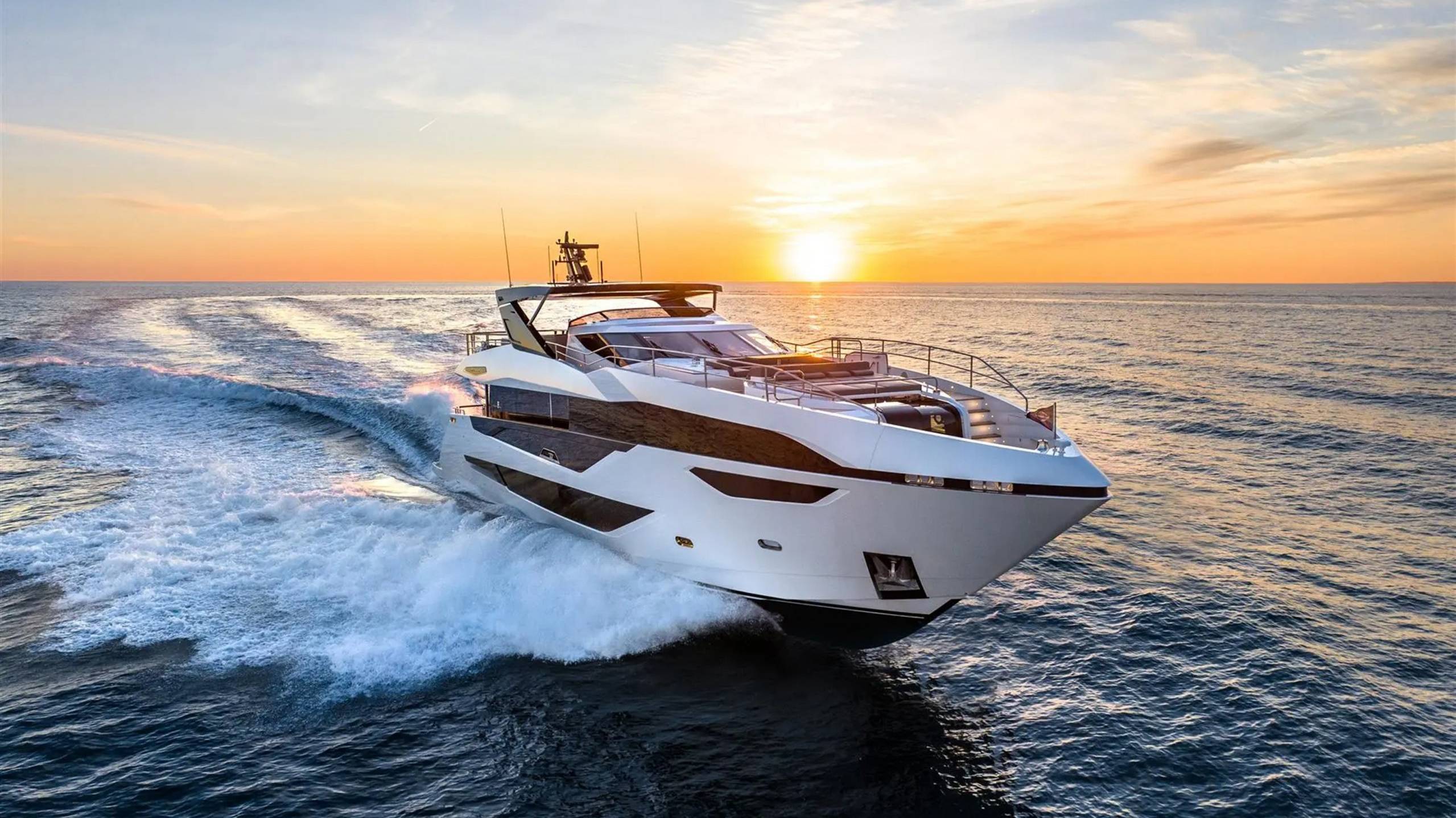 A sleek white yacht cuts through the ocean waves at sunset, leaving a foamy wake under a warm, golden sky.