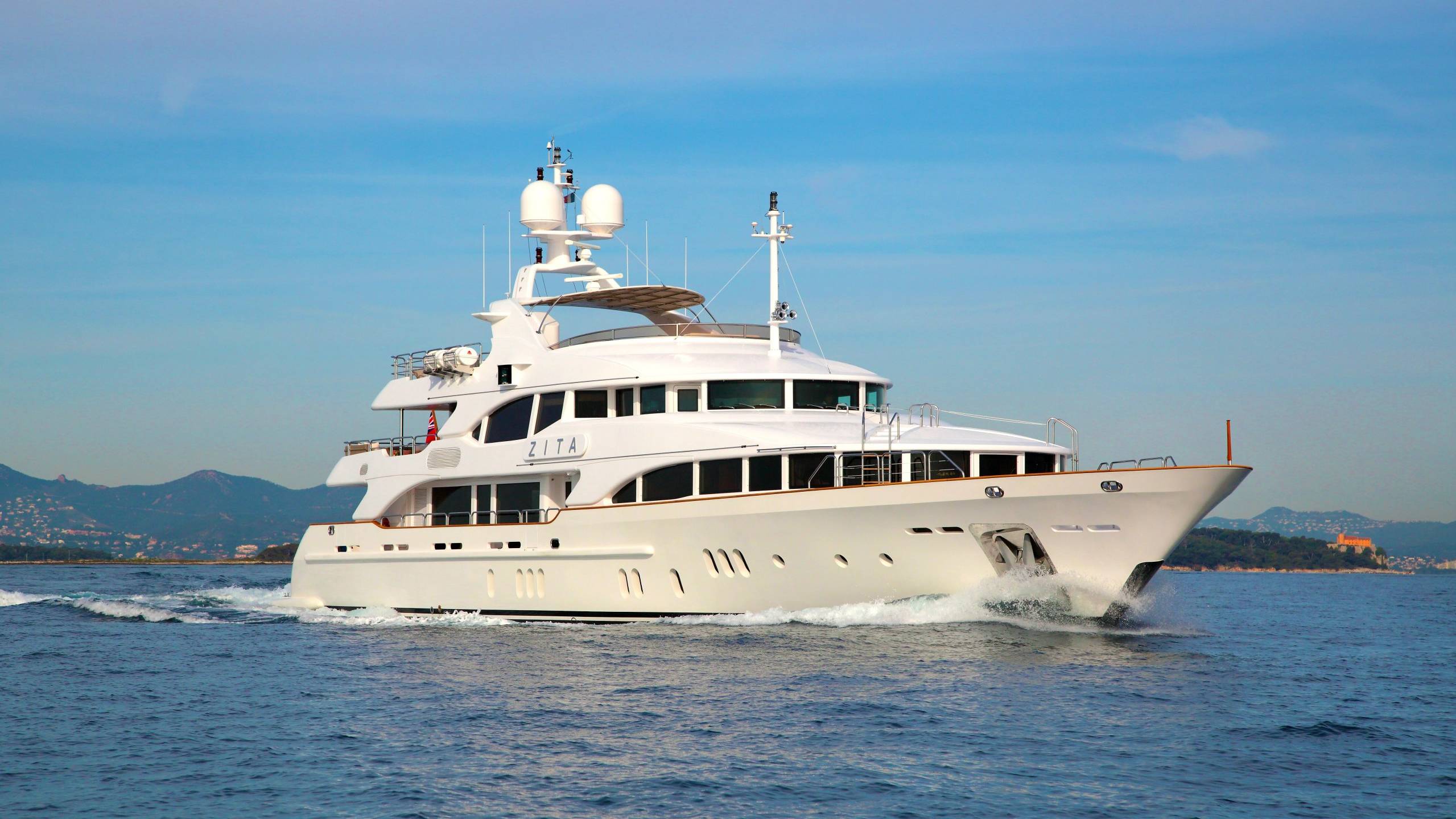 The gleaming white yacht ZITA cuts through the azure waters, with a picturesque coastline and mountains in the background.