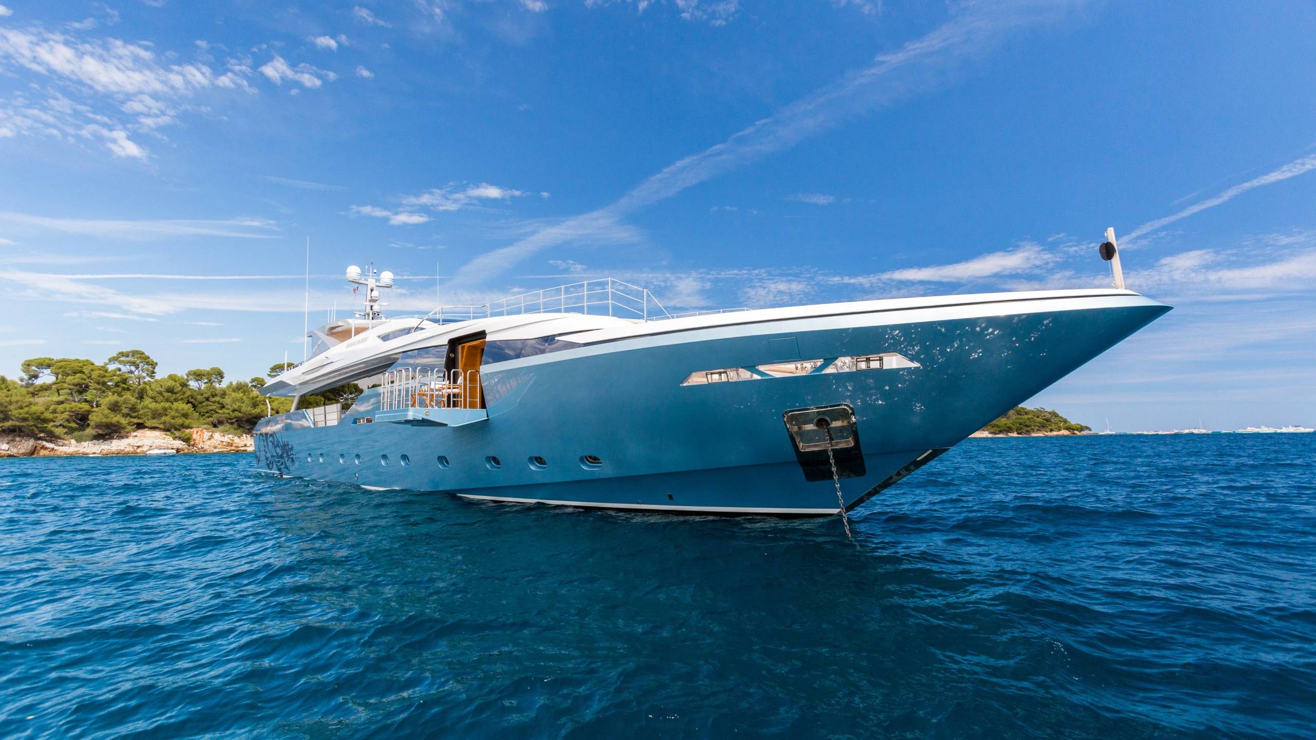 A large, luxurious yacht in a vibrant blue color floats on the calm, azure waters of a scenic coastal area surrounded by lush greenery.