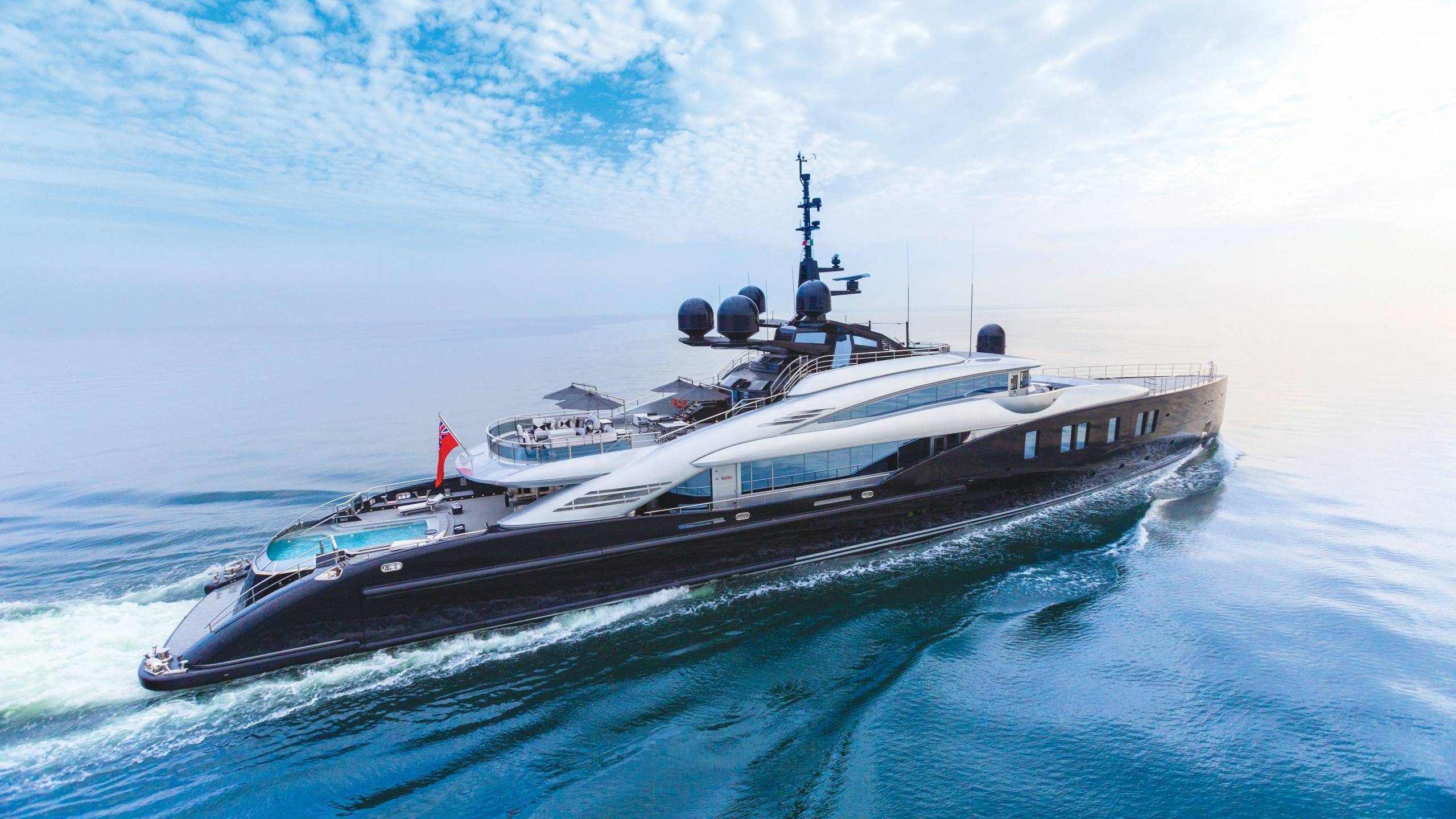A sleek, modern superyacht glides through calm blue waters under a sky dotted with fluffy clouds.