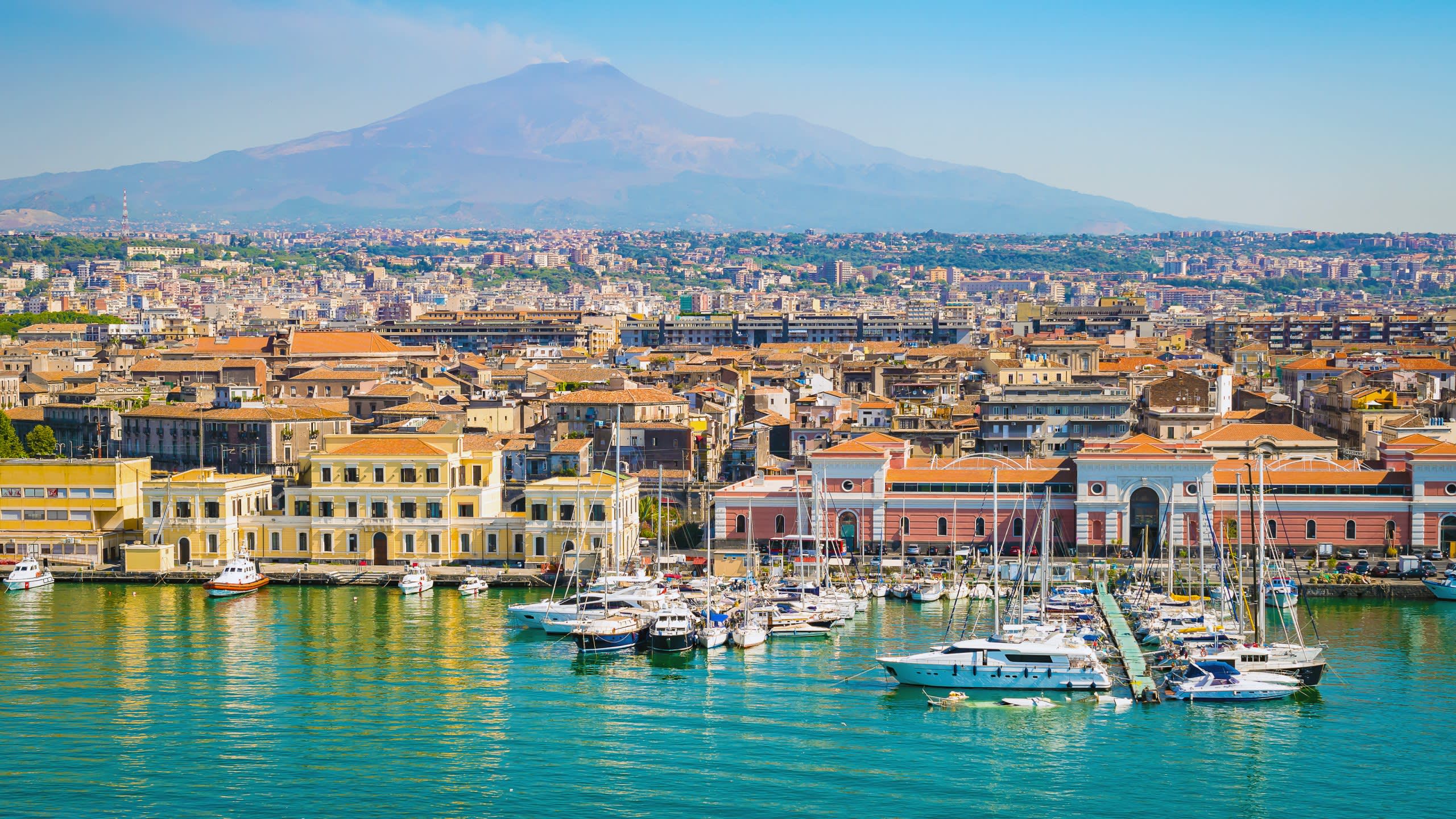 A vibrant harbor filled with boats is nestled below a sprawling city with a majestic volcano in the distance.